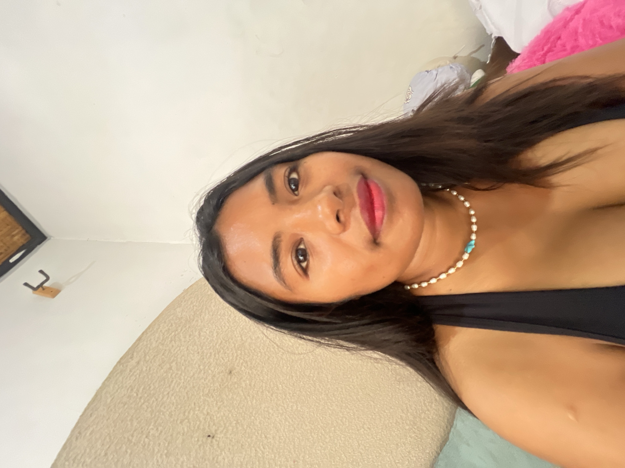 A young woman with long, dark hair and brown eyes is taking a selfie indoors. She is wearing a black halter top and a pearl necklace with turquoise accents. Her lips are painted red. Behind her, a textured, light-colored wall curves upwards, and a white wall is visible to the right. A wooden hook is mounted on the white wall. The scene appears to be in Denpasar, Indonesia.