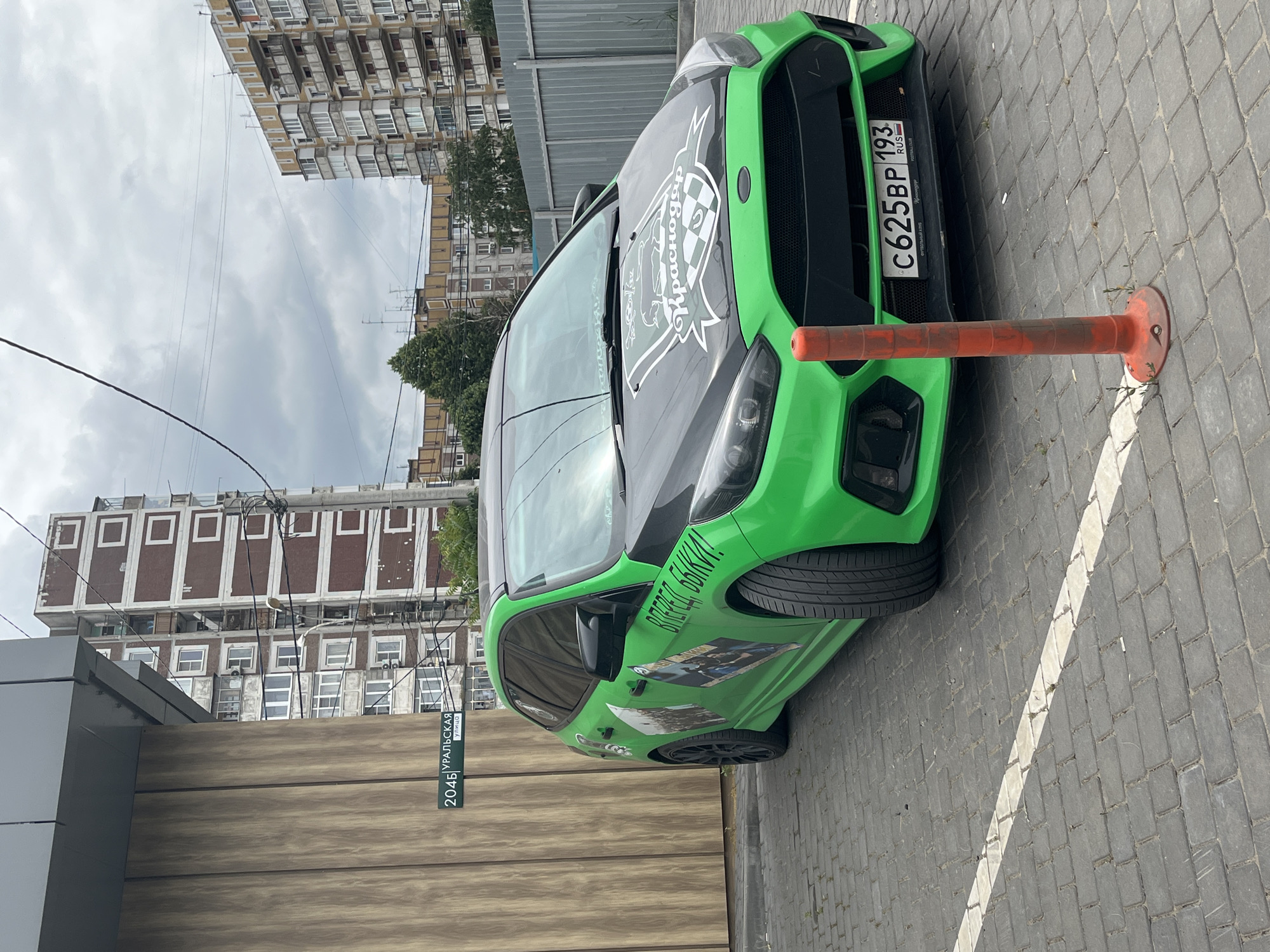 A bright green Ford Focus ST is parked diagonally in a paved lot in front of a building with a wooden facade. The car has a black hood and roof, and features numerous stickers. A prominent sticker on the hood displays a crest with the word "Krasnodar" in stylized script. The car's license plate reads "C625BP 193 RUS". On the side of the car, there are several rectangular stickers depicting images, and text that appears to be "ВПЕРЁД БЫКИ!". An orange traffic cone stands in front of the car. In the background, apartment buildings typical of urban Russian architecture are visible under a cloudy sky. A street sign with the text "204Б УРАЛЬСКАЯ УЛИЦА" is mounted on the wooden facade. The scene is a street-level view in Krasnodar, Russia.