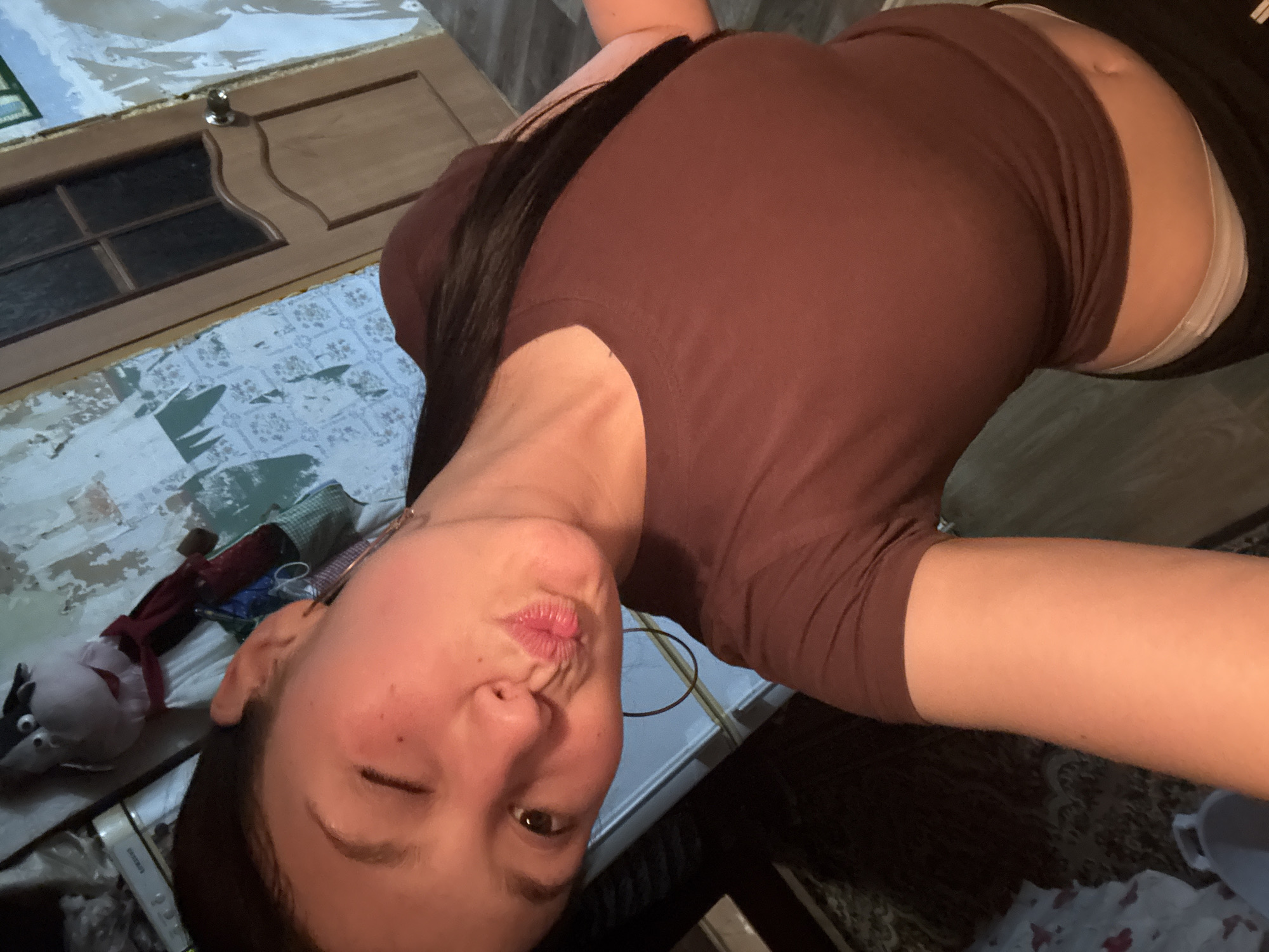 A woman with dark hair and brown eyes is posing for a selfie in an indoor setting. She is wearing a brown, short-sleeved shirt and what appears to be dark shorts or pants, with a glimpse of white underwear showing above the waistband. Her face is tilted towards the camera, and she is winking one eye and puckering her lips in a kissing gesture. The location is not recognizable as the city of Фоки, Russia, based on the visible elements in the image. To the left of the woman, a patterned fabric or wallpaper is visible, and there are some small objects and what looks like a stuffed animal. Part of a wooden door is also visible in the upper left corner. The overall lighting in the room appears to be dim.