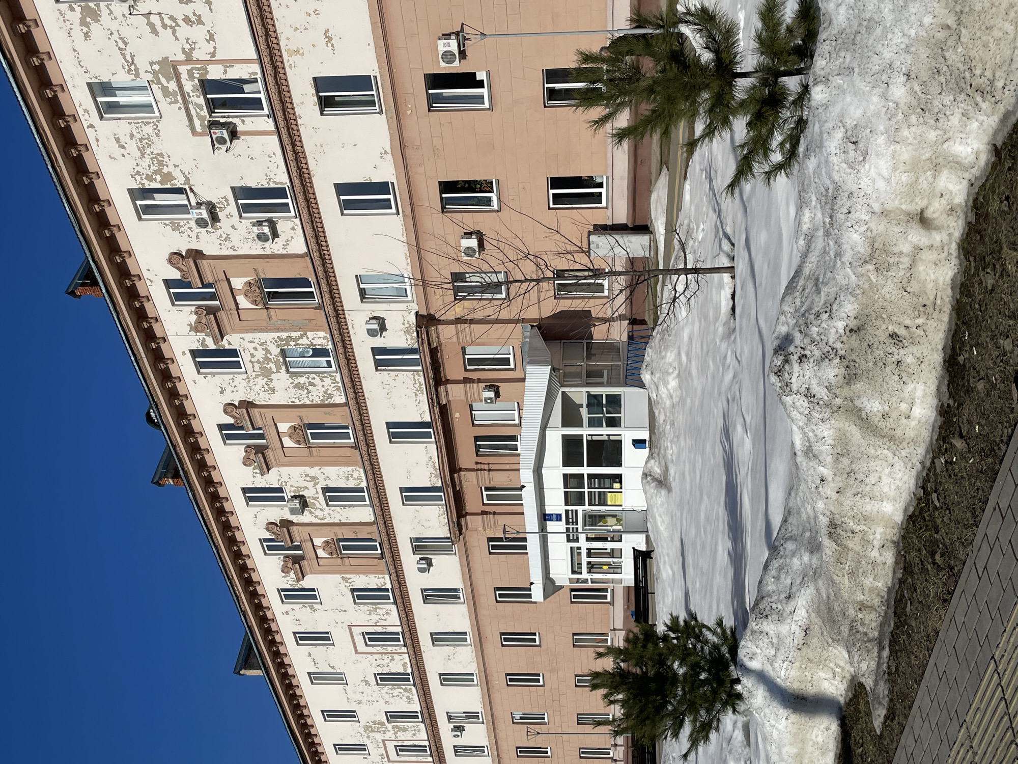 The image shows the exterior of a building in Izhevsk, Russia, likely in late winter or early spring based on the presence of snow. The building is multi-story with a mix of light pink and beige colored facade sections. Many windows are visible, with some containing air conditioning units. The building is partially worn, with peeling paint visible on some sections. There's a glass-enclosed entrance area visible near the center of the frame. Snowbanks are adjacent to the building. Two small trees are on the building's side. The sky is clear blue.