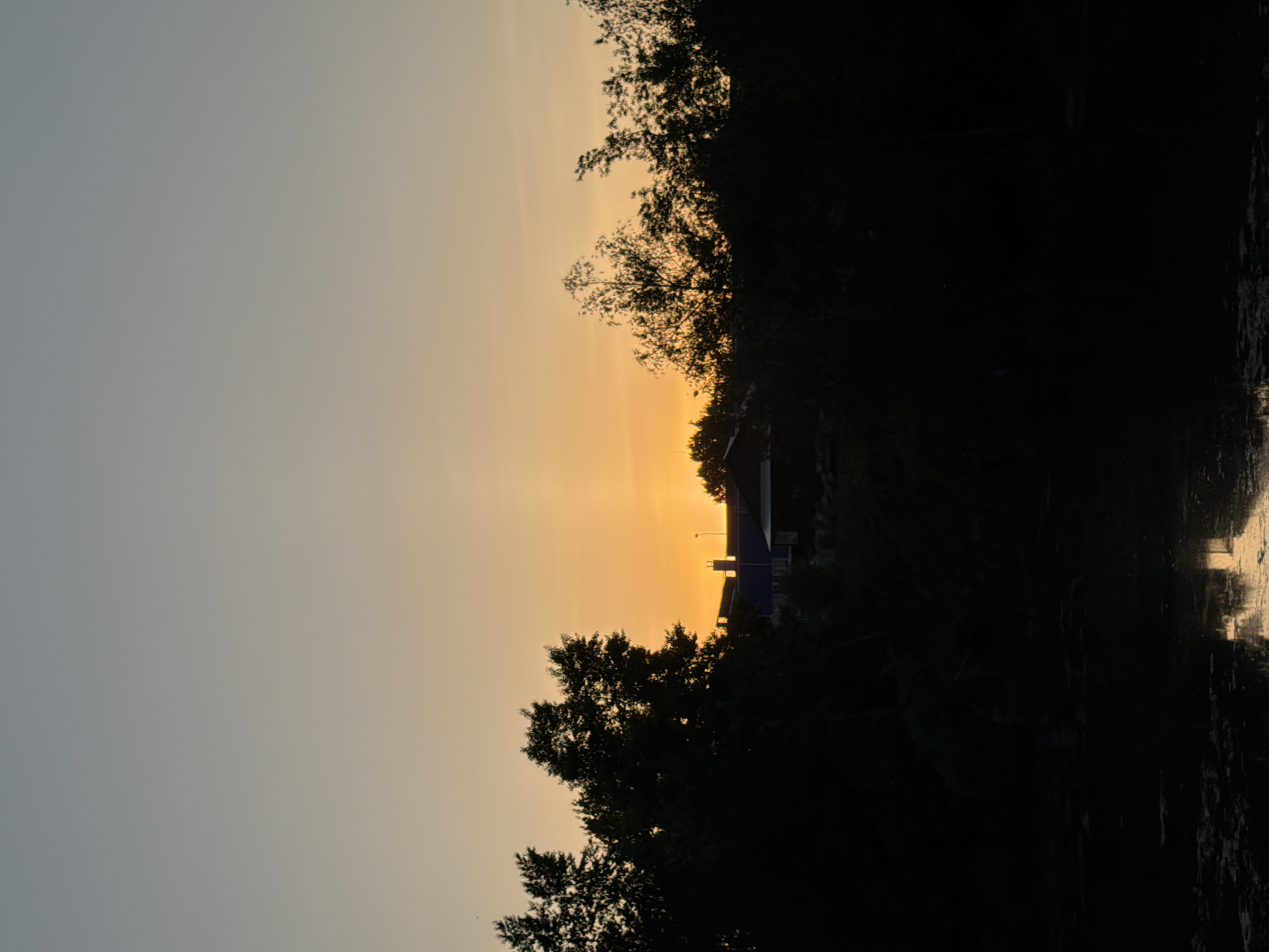 The image shows a sunset scene in Chaikovsky, Russia. The sun is setting, casting a golden glow in the sky. The horizon is partially obscured by trees and a building. A body of water reflects the light from the sky on the right side of the image. The left side of the image shows a blue sky.
