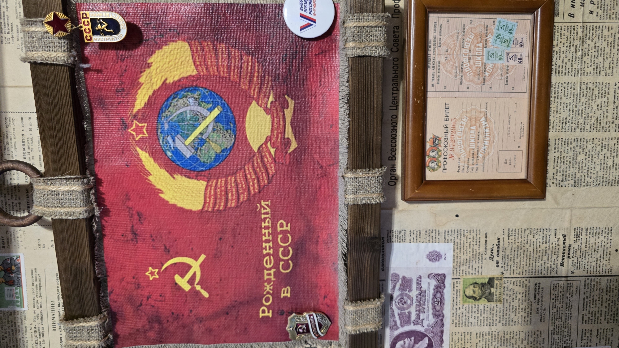 The image displays a Soviet-era banner featuring the hammer and sickle symbol and the text "Рожденный в СССР" (Born in the USSR). Below the banner, a framed document, likely a Soviet Union trade union membership card, is visible. The background consists of vintage newspapers and what appears to be a wooden display or frame. The overall presentation suggests a historical or nostalgic display, possibly in a personal collection or a small museum. The location in the city of Верхнеказымский, Russia, is not directly indicated by any visual elements within the provided image.