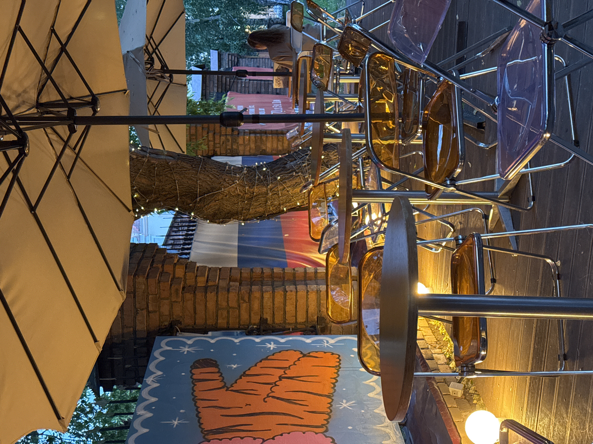 The image shows an outdoor seating area at an establishment in Krasnoyarsk, Russia. The seating area is comprised of several round tables with metal legs and wooden tops, surrounded by transparent plastic chairs with metal frames. Two large patio umbrellas, one light-colored and one darker, provide shade over the seating. The ground is a wooden deck.
To the left, a wall features a colorful mural of an orange tiger-like creature with a blue background, adorned with white stars. Adjacent to the mural is a brick pillar, and behind it, a large Russian flag is visible, hanging vertically and partially obscured by a tree. The tree itself is wrapped with strings of small, white lights.
In the background, on the right side of the image, a woman with long dark hair pulled back in a ponytail is seated at a table, facing away from the camera. She is wearing a light-colored top. Another sign, partially visible behind her, has Cyrillic text and the word "epos". The overall ambiance suggests an evening setting, with artificial lighting casting warm glows on the scene. The presence of the Russian flag and the Cyrillic text strongly indicate the location as Krasnoyarsk, Russia.