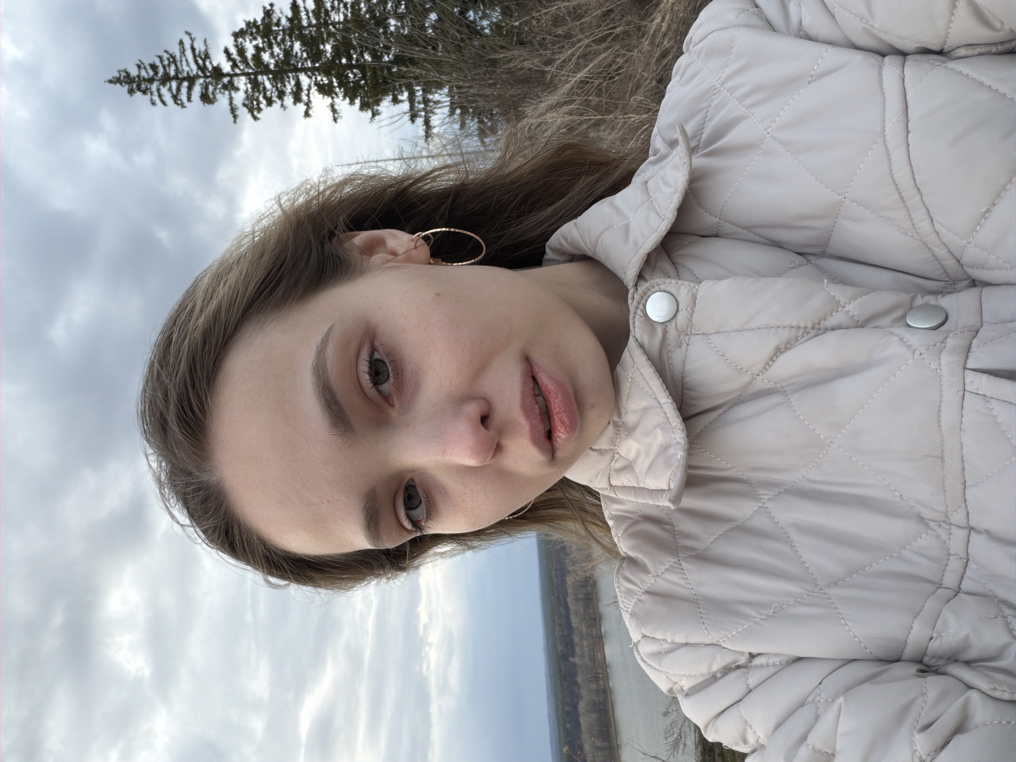 A woman is taking a selfie in an outdoor setting in Chaykovsky, Russia. She is wearing a quilted jacket and gold hoop earrings. Behind her is a natural scene with bare trees and a cloudy sky. A river or a large body of water is also visible in the background.