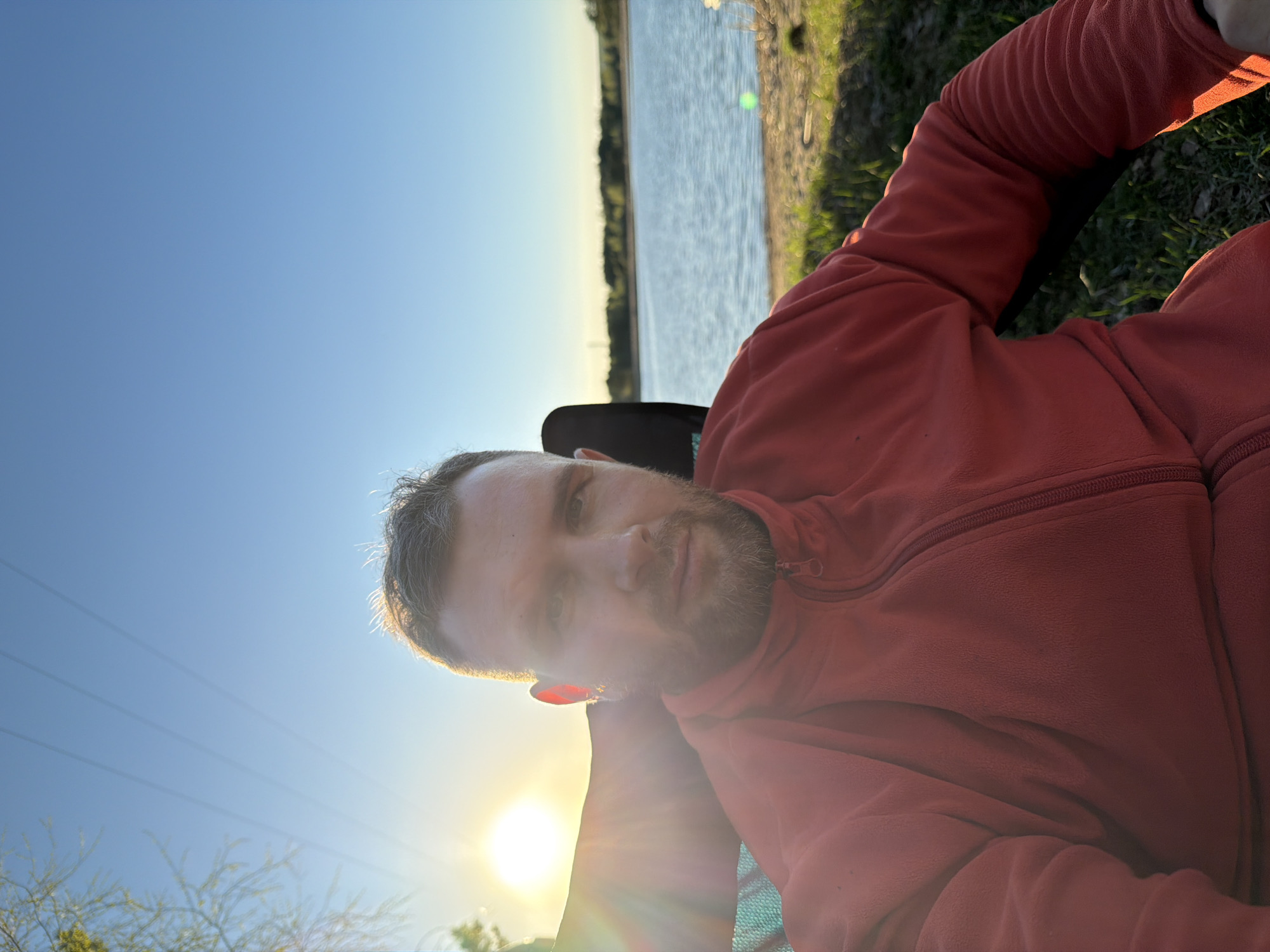 A man with short, light-brown hair and a beard is lying down in a chair, wearing a red fleece jacket. He is positioned outdoors, as evidenced by the presence of grass, water (likely a lake or river), and trees in the background. The sky is blue and clear, with the sun shining brightly, creating a lens flare effect. The location is possibly near water, which is consistent with the city of Chaykovsky, Russia, known for its proximity to the Kama River and Votkinsk Reservoir.