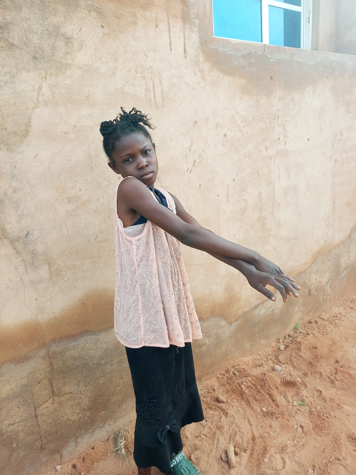 A young Black girl with braided hair stands in front of a textured, beige wall with a blue-paned window at the top. She wears a light pink, sleeveless, sequined top over a dark bra and a black skirt with decorative accents and a bow. Her arms are extended forward, one slightly bent and resting on the other. Her hands are open and facing forward. The ground in front of her is sandy and appears to be outdoors.