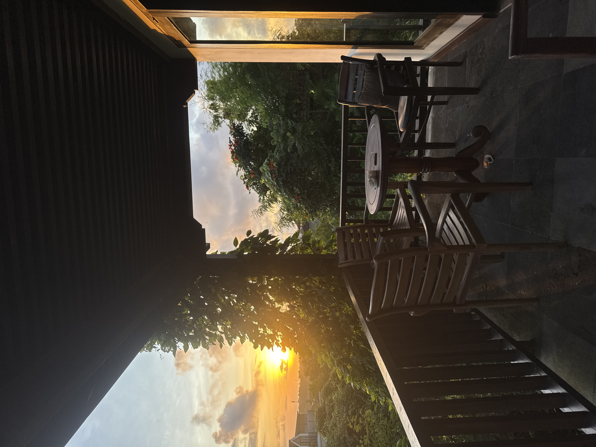 The image shows a sunset view from a balcony in Ungasan, Indonesia. The balcony has wooden furniture, including a round table and several chairs. Lush tropical vegetation is visible beyond the balcony railing, and the setting sun is partially obscured by trees. The sky displays orange and yellow hues. The balcony floor is dark stone. A building roof can be seen on the lower right side of the balcony.