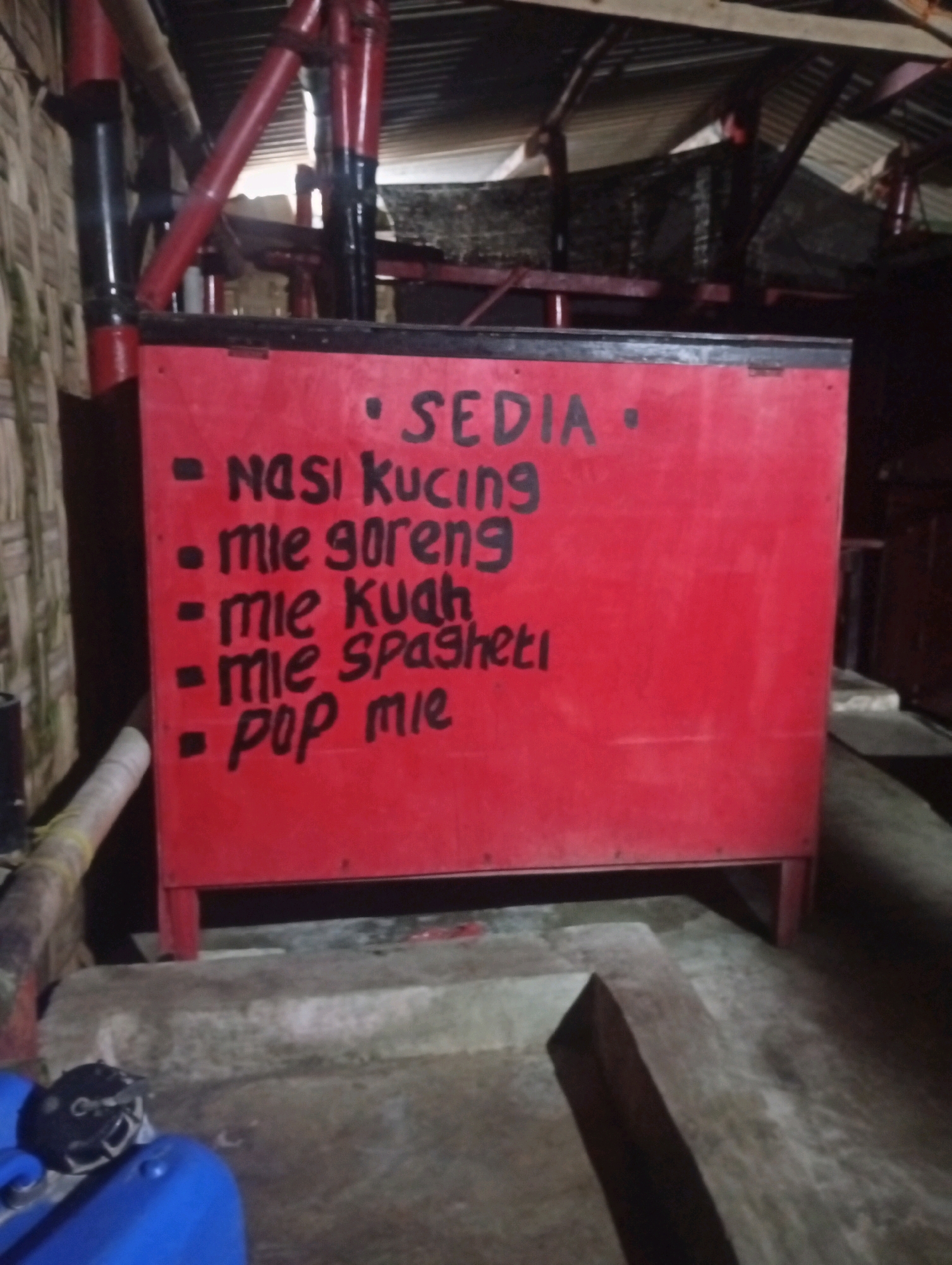 The image shows a red metal food stall with a list of menu items written in black marker. The stall is located outdoors in what appears to be a rural or semi-urban setting in Banowan, Indonesia. The menu lists: "SEDIA" (meaning "available" or "offering"), followed by "NASI KUCING" (cat rice), "mie goreng" (fried noodles), "mie kuah" (noodle soup), "mie spaghetti", and "pop mie". The stall is supported by red metal legs and is situated on a concrete base. In the background, there are corrugated metal roofs and bamboo walls, suggesting a rustic or simple construction. The lighting is dim, indicating it might be evening or the area is poorly lit. A blue plastic container and a black object are visible in the lower left corner of the image, and a wooden beam lies on the ground in the foreground. There are no people visible in the image.