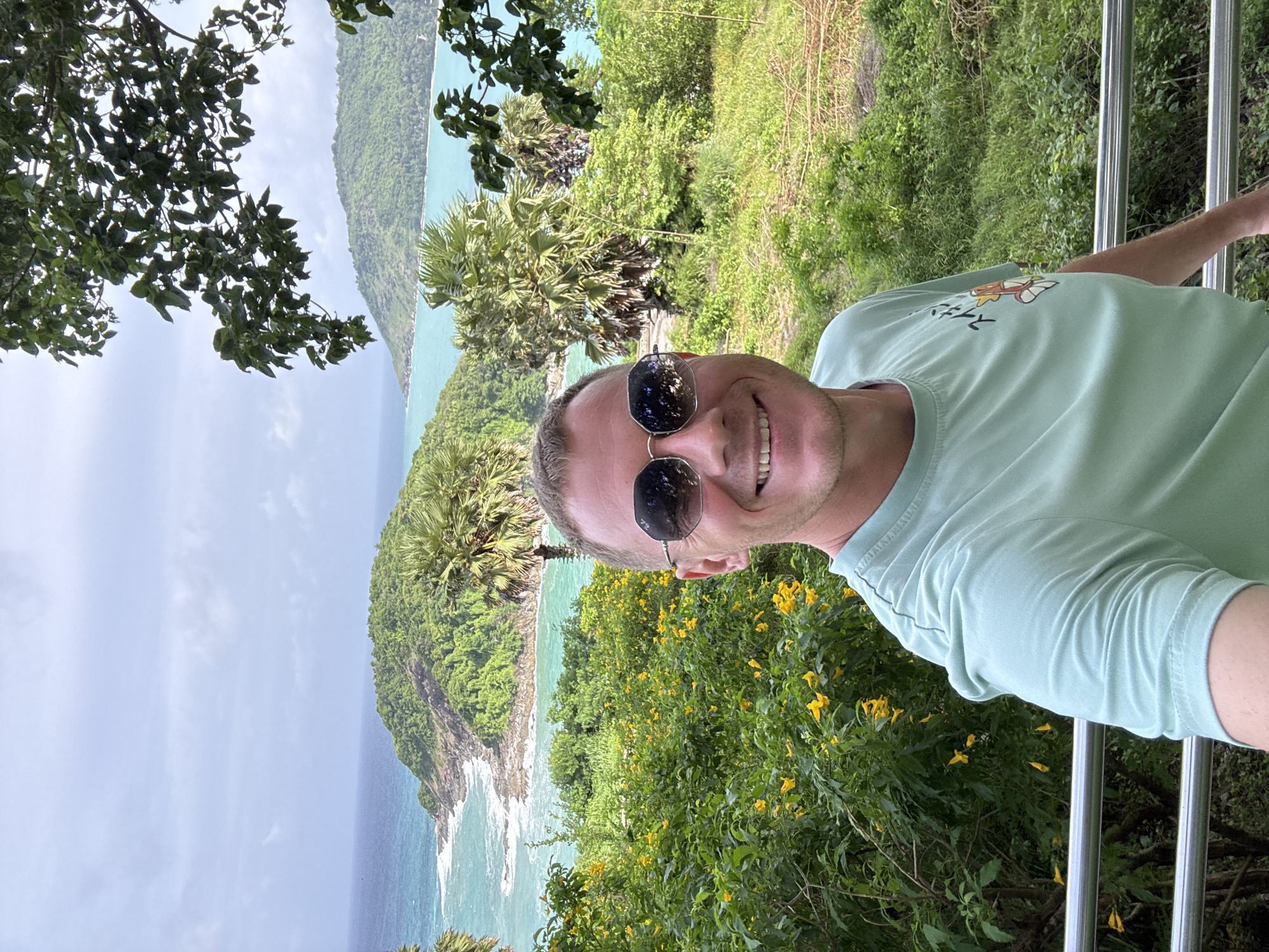An outdoor, daytime, eye-level selfie is captured from the chest up of a smiling, fair-skinned man wearing sunglasses and a light green t-shirt. He is positioned in the foreground, with lush green foliage and yellow flowers surrounding him. In the background, a turquoise ocean with white-capped waves meets a series of verdant, tropical islands. Palm trees are visible among the vegetation. The scene is likely located at a viewpoint overlooking the Andaman Sea in Rawai, Thailand.