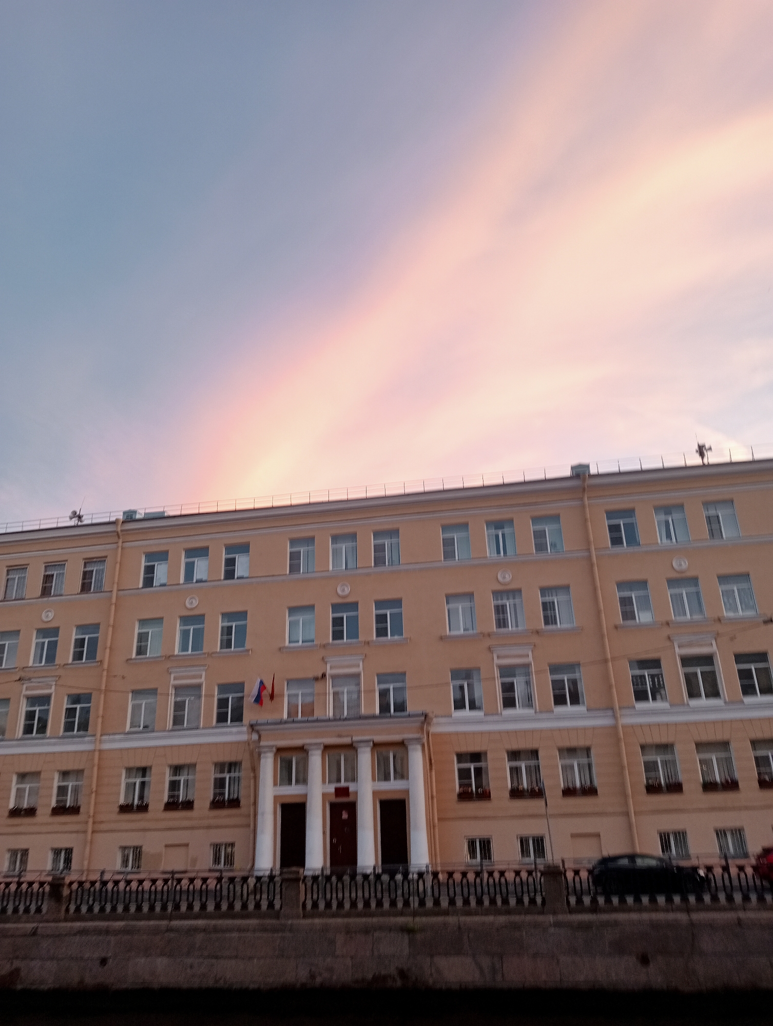 The image shows a large, multi-story building with a yellow facade, likely a school or administrative building, situated in Saint Petersburg, Russia. The building has numerous windows with white frames and a classical architectural style. In front of the building, there is a canal or river with a decorative black metal fence along the embankment. Several cars are parked along the street in front of the fence. The sky above is a soft gradient of blue and pink, suggesting either sunrise or sunset. Two flags, one appearing to be the Russian flag, are displayed on the building. The overall scene captures an urban landscape with a touch of natural beauty from the sky.