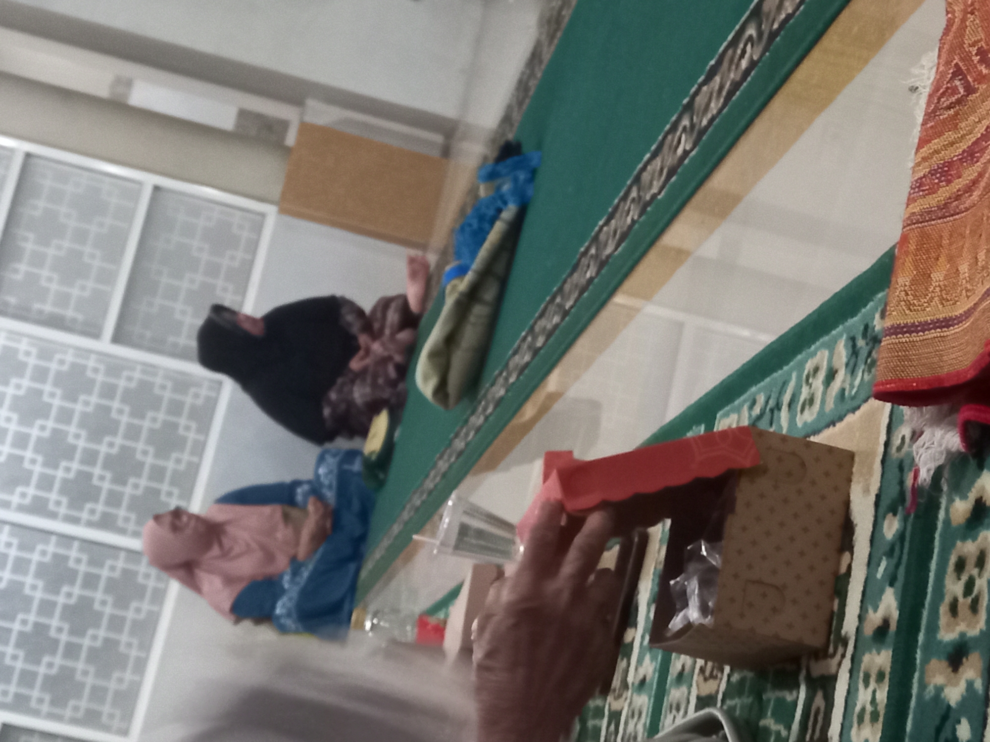 In the city of Banyumulek, Indonesia, two individuals are seated on a green carpet in what appears to be an indoor gathering. The individual on the left wears a light pink hijab and a blue garment, while the individual on the right is dressed in a black hijab and patterned clothing. In the foreground, a person's hands are visible interacting with a small, brown, patterned box with a red lid. Nearby, a clear plastic cup with a straw and a bottle with a red liquid are present on a table. The background shows a wall with a grid-like pattern, suggesting an interior setting. The overall scene conveys a sense of a casual, possibly religious or social, gathering.