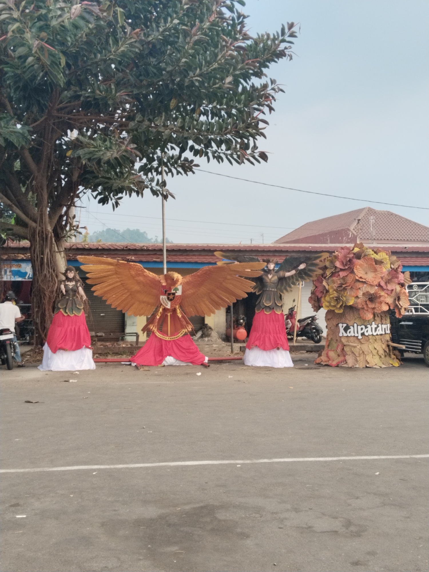 In Tawangmangu, Indonesia, a street scene features several large, theatrical figures. On the left, a person on a motorcycle is visible. To the right of the motorcycle, a life-sized doll figure stands dressed in a dark, layered bodice and a long, flowing red skirt over a white base. Next to this figure is a large, golden eagle effigy with outstretched wings, also adorned with red and white fabric at its base. Further to the right, another doll figure, similarly dressed, is positioned, its arms outstretched. The fourth prominent object is a large, stylized tree effigy with layered, leaf-like decorations in autumnal colors, bearing the word "Kalpataru" in white lettering. Behind these figures, buildings with reddish-brown tiled roofs are visible, as well as a black pickup truck. The ground is paved asphalt, with some debris scattered. A large tree with dense foliage stands to the left, its roots exposed. The sky is a pale, overcast blue.