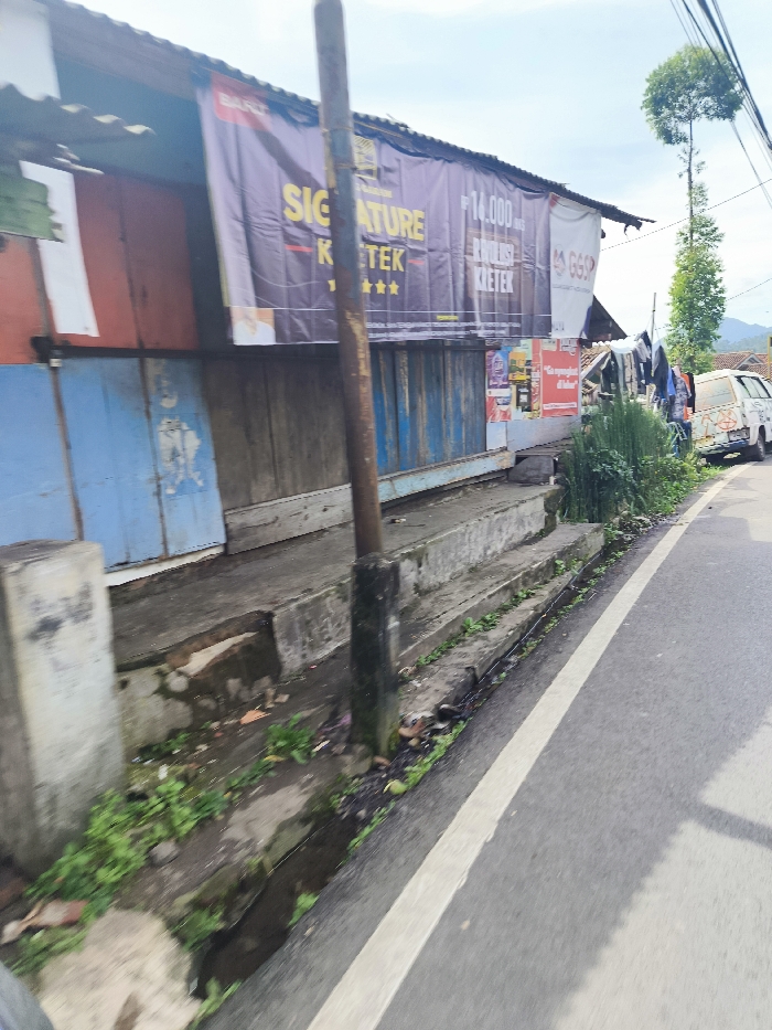 The image shows a street scene in Rancabali, Indonesia (latitude: -7.123, longitude: 107.431). A small, single-story building with wooden siding and a corrugated metal roof is on the left side of the image. The building has several large signs on its front, including one that says "Signature Keter" and another that says "Rp. 14.000 Brushless Keter". The building has a blue door and a small, concrete step leading to the door. There is a large, rusty metal post in front of the building. There is a small, white van parked on the side of the road next to the building. There is also a green, tree in the background. The street is paved with asphalt. There is a white painted line in the middle of the road. There is a narrow, grassy area between the building and the road, with small plants growing in it. There is a shallow, open drainage ditch beside the road. The image is taken from a low angle, and it looks like the photographer is standing on the edge of the road, facing the building. The image is in focus, and it is well-lit.