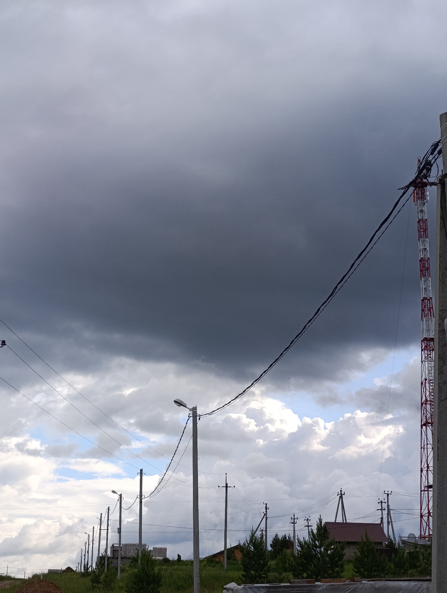 The image depicts a wide shot of a rural or suburban area under a dramatic, overcast sky. In the foreground, power lines stretch across the frame, supported by numerous utility poles. A tall, red and white communications tower is visible on the right side of the image. In the middle ground, a row of houses and partially constructed buildings are scattered amongst green trees and grass. The overall atmosphere is somber due to the dark, stormy clouds dominating the sky. The scene appears to be located in Chaikovsky, Russia, given the architectural style of the houses and the presence of utility infrastructure commonly found in such regions. There are no people visible in the image.