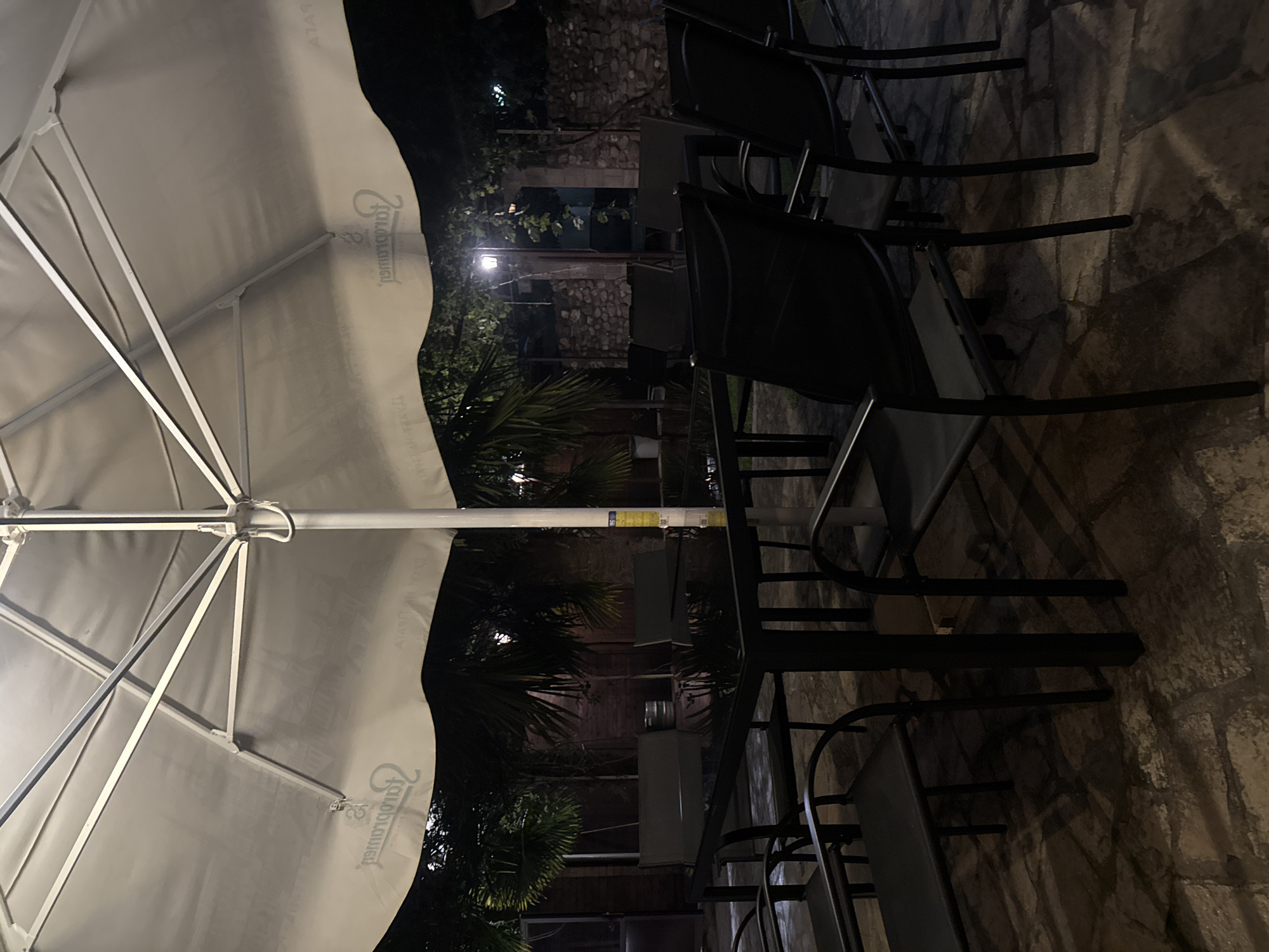 The image shows an outdoor seating area at night, likely a restaurant or cafe, located in Dimitrovgrad, Bulgaria. A large white umbrella covers a black rectangular table surrounded by several black chairs. The umbrella has a logo printed on it. The ground is paved with irregular stones. In the background, there is foliage, a stone wall, and a lamppost. The scene appears to be dimly lit. There are no people visible in the image.