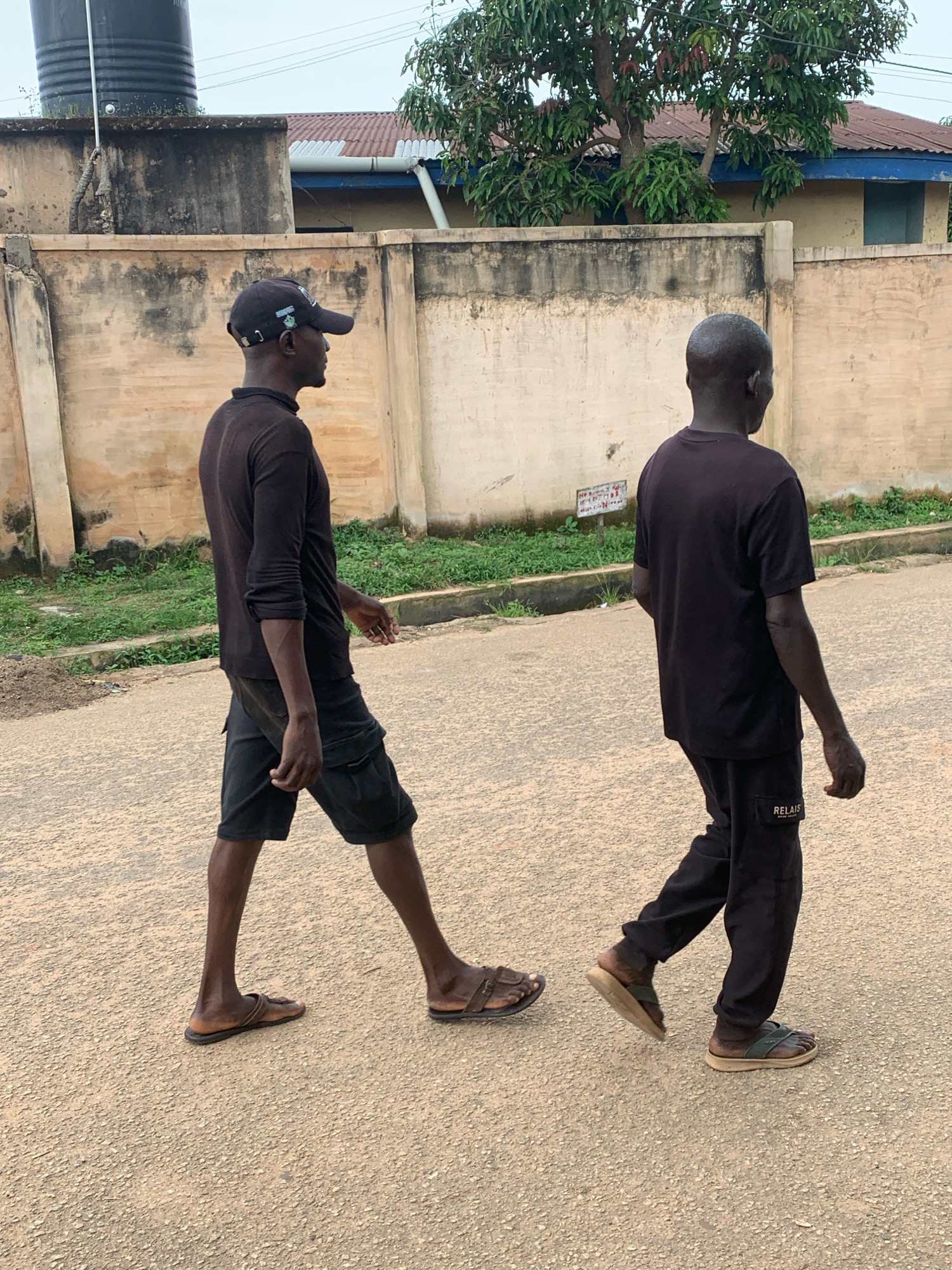 Two men are walking on a paved road. The man on the left is wearing a black long-sleeved shirt, black shorts, and a black baseball cap. The man on the right is wearing a black t-shirt and black pants. They are walking in the same direction. The environment appears to be a residential area with buildings and vegetation in the background. The sky is overcast. This scene is located in the city of Jos, Nigeria.