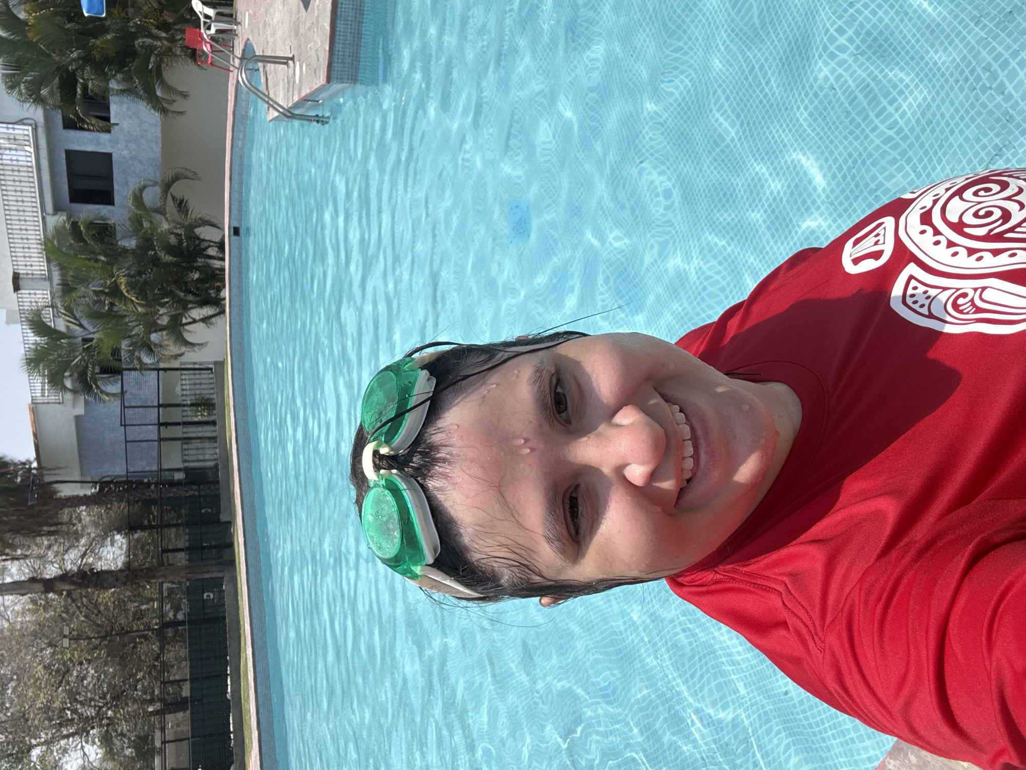 The image depicts a smiling person taking a selfie in a vibrant blue swimming pool under bright daylight in Tres de Mayo, Mexico. The subject, positioned in the foreground, is partially submerged in the water, with their head and upper torso visible. They have wet, dark hair and water droplets on their face, suggesting recent activity in the pool. Green swimming goggles are pushed up onto their forehead. The person is wearing a red long-sleeved shirt adorned with distinctive white tribal-like patterns on the right shoulder and arm. Their wide smile reveals their teeth, conveying a cheerful and happy emotion.
The setting is an outdoor swimming pool, characterized by clear, light blue water reflecting sunlight, with a visible grid pattern from the tiles at the bottom. A light-colored concrete or tiled deck surrounds the pool, where a metallic ladder for access can be seen, along with red and white plastic chairs further back. The background features lush green foliage, including palm trees, partially obscuring a multi-story building with light-colored walls and dark windows. A white fence or railing is also visible, indicating a residential or recreational environment. The bright, clear conditions suggest sunny weather. No discernible text is present, apart from the decorative patterns on the shirt.