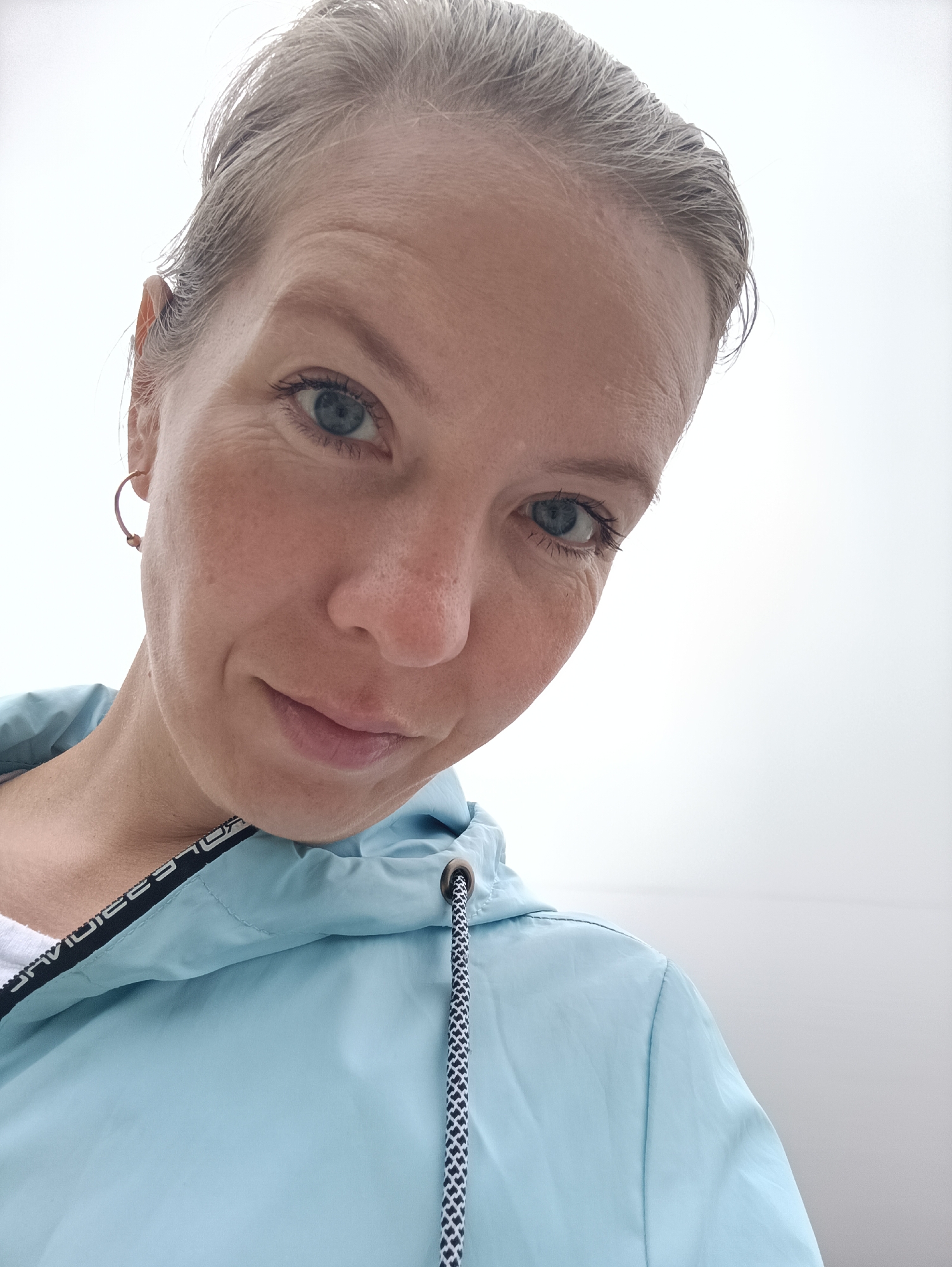 A close-up, upward-angled selfie features a person, likely female, against a plain, bright white background, which could be an overcast sky or a very bright indoor setting. The subject has light skin, blue eyes, and light brown hair with visible grey strands, pulled back from her face. She has a calm expression with a subtle, gentle smile, looking directly into the camera.
She is wearing a light blue, hooded jacket over a white inner garment. The jacket features a black and white checkered drawstring for the hood and a black trim along the neckline. Text printed in white on this black trim is partially visible, spelling out "DAVIDDES" followed by obscured letters that suggest a brand name like "DAVIDDESPORT" or similar sportswear. A small, golden-colored hoop earring is visible in her left ear.
The bright, diffuse lighting indicates daytime. The white background offers no specific environmental cues beyond the subject. The image was captured in Chaikovsky, Russia.