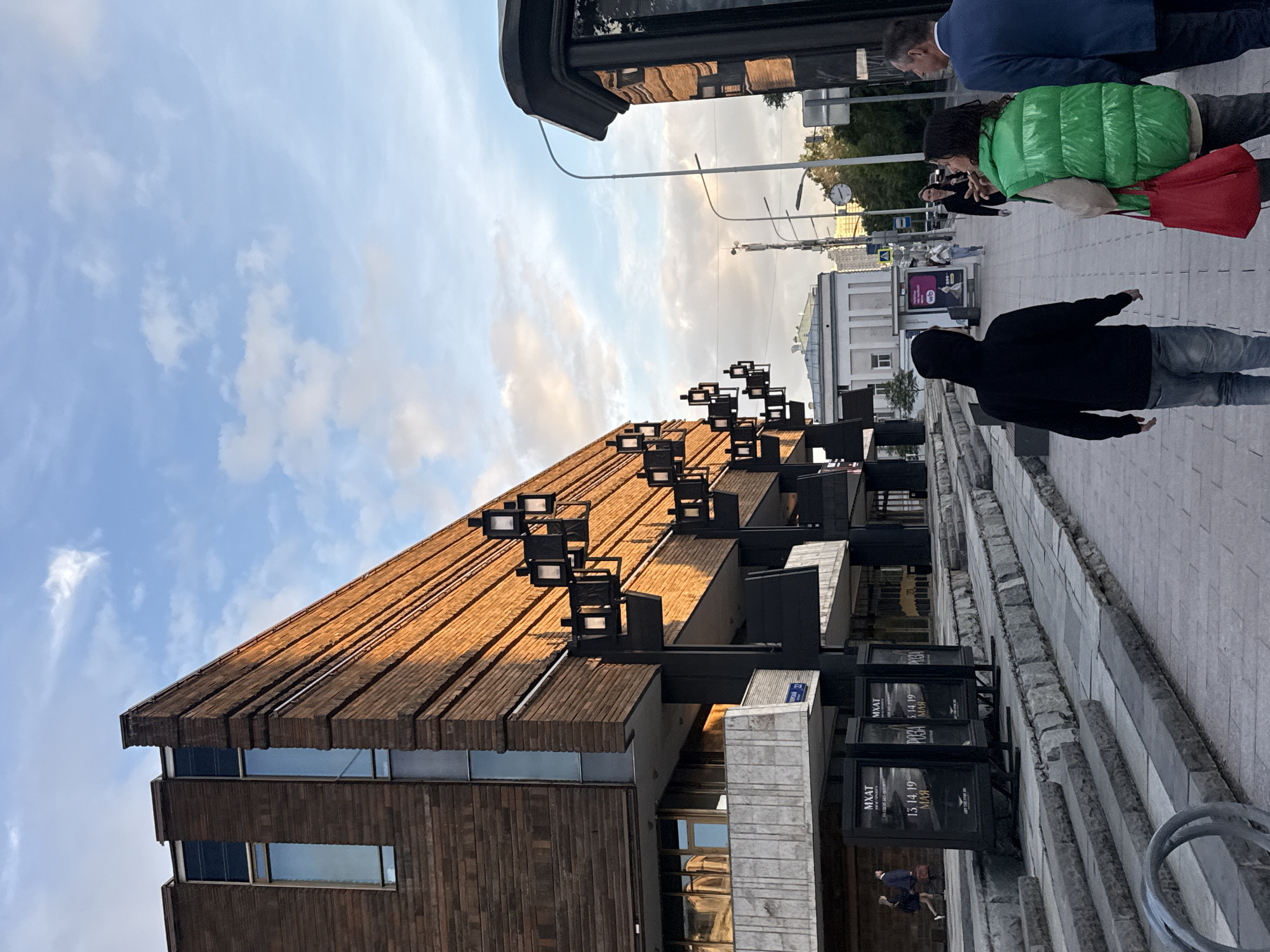 The image captures a vibrant urban street scene, likely in Moscow, during the late afternoon or early evening, illuminated by warm sunlight under a partly cloudy sky.
The most prominent subject is a large, multi-story building on the left, characterized by its distinctive brown, textured facade, possibly constructed from brick or wood panels. The upper section features a slatted or stepped roof design, adorned with multiple cube-shaped light fixtures mounted on dark beams. Several windows reflect the sky. The building is identified by the text "MXAT" (Moscow Art Theatre) visible on signs and on its structure, confirming the location context.
A wide, paved sidewalk runs alongside the building on the right. Several people are visible walking along it. In the foreground, a person in a black hooded sweatshirt and blue jeans walks away from the viewer. Further back, a person in a bright green puffer jacket, carrying a red bag, walks towards the camera alongside another individual in a dark suit. Other pedestrians are scattered further down the street.
Along the sidewalk, multiple black display boards are arranged, prominently featuring the text "MXAT" and the dates "13.14.19 МАЯ" (May). In the background, streetlights, utility poles, and a street clock are visible. The sky is light blue with scattered white, wispy clouds. The scene conveys a sense of calm, everyday activity as people move about in front of a significant architectural landmark.