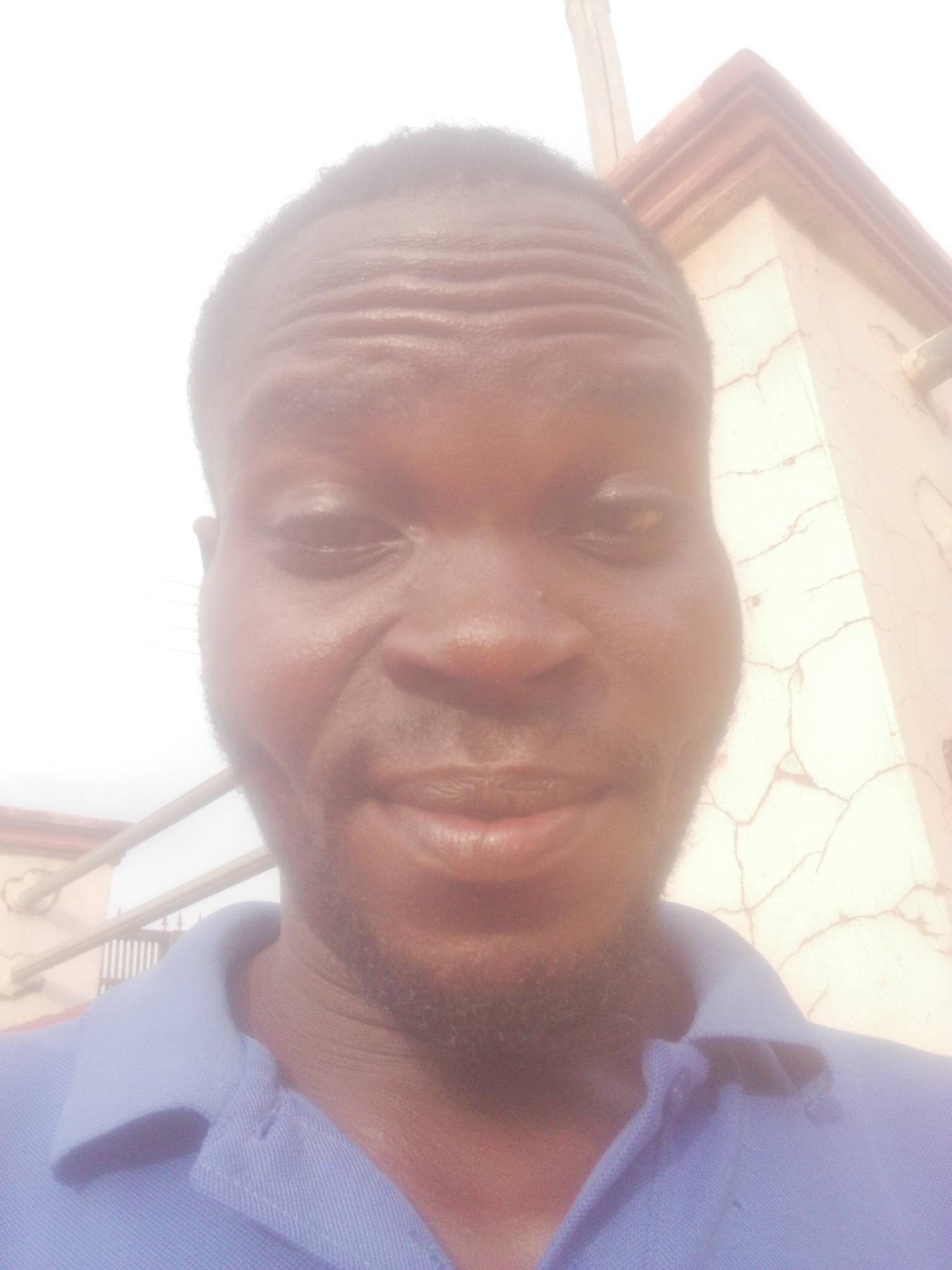 A man is shown in a close-up selfie. He has dark skin, a beard, and is wearing a blue collared shirt. Behind the man is a beige building, possibly located in a city.