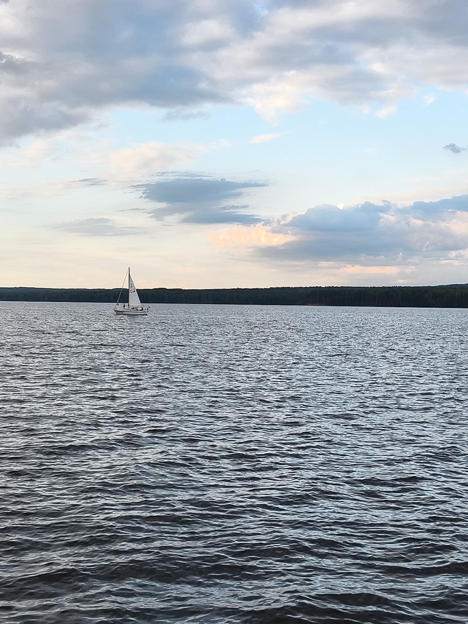 A sailboat with a white sail glides across a calm lake. The boat is positioned to the left of the image's center. A dark, dense forest line stretches across the horizon behind the lake, separating it from the sky. The sky is filled with a mixture of clouds, with areas of pale blue peeking through. The image appears to be taken in Chaikovsky, Russia, given the distinctive features of the landscape and common recreational activities in the region.