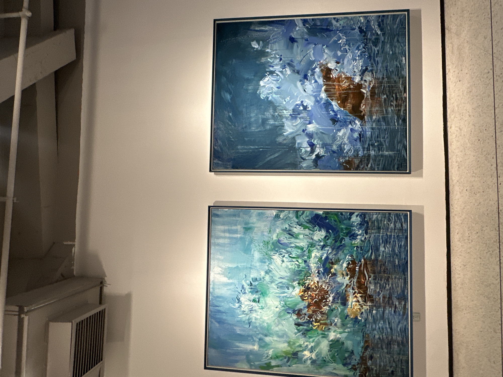 Two framed paintings hang on a white wall. The top painting features shades of blue and white with a patch of brown. The bottom painting also uses blue and white, but incorporates more green and a similar brown patch. To the left of the paintings, there is a white structure with pipes and a radiator. To the right, there is a wall with vertical lines. The location in Banan, China is not specified.