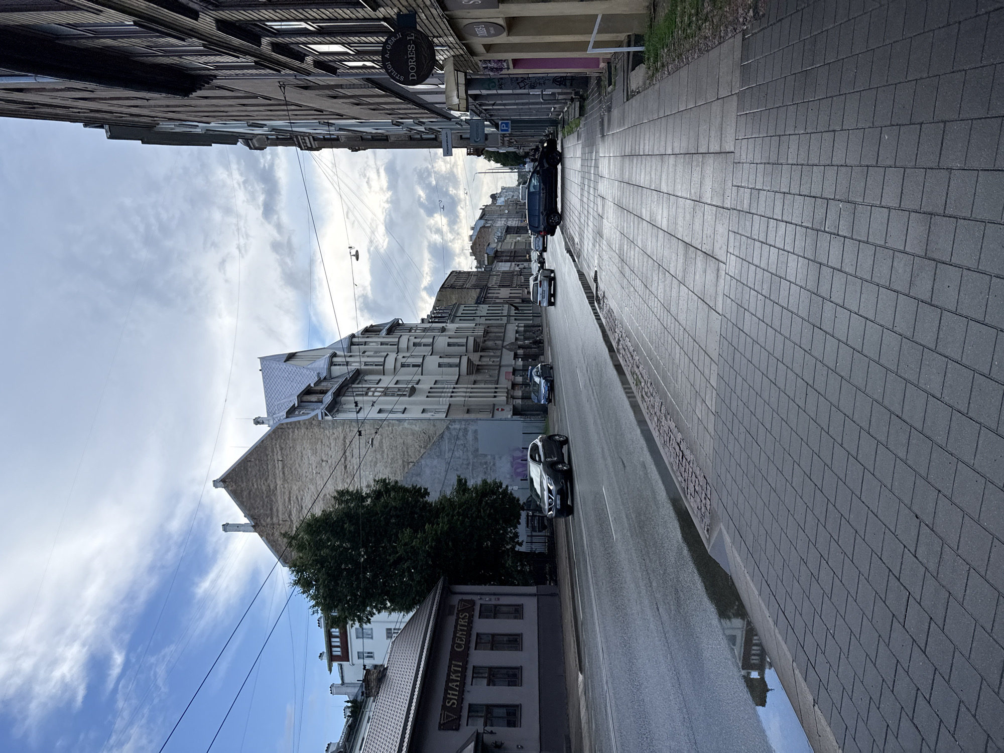The image depicts a street scene in Riga, Latvia, under an overcast sky. The perspective is from a sidewalk, looking down a moderately busy street lined with buildings. On the left side of the street, a low-rise building with a sign that reads "SHAKTI CENTRS" is visible. Further down the street on the left, taller, older buildings with distinctive architecture dominate the skyline. On the right side, a row of multi-story buildings with numerous windows and some signage is present, including a sign for "DORES-L." Several cars are on the road, some in motion and others parked. A large puddle is visible on the wet street surface near the curb on the left. The sidewalk is paved with rectangular gray tiles. The overall impression is one of an urban environment, possibly in the older part of the city, with a mix of architectural styles.