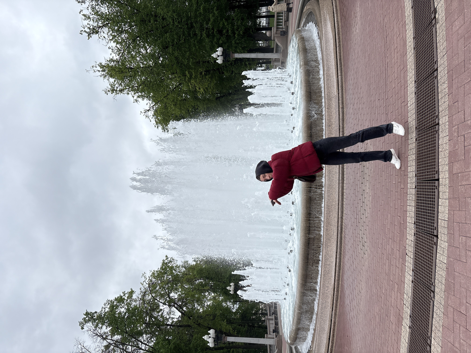 The photo captures a person in motion at a fountain in Minsk, Belarus. The individual is wearing a dark beanie, a maroon puffer jacket, black pants, and white shoes. They are positioned along the edge of a circular fountain that is actively spraying water. Green trees and lampposts are visible behind the fountain. The foreground shows a brick-paved area and a metal grate. The sky is overcast.