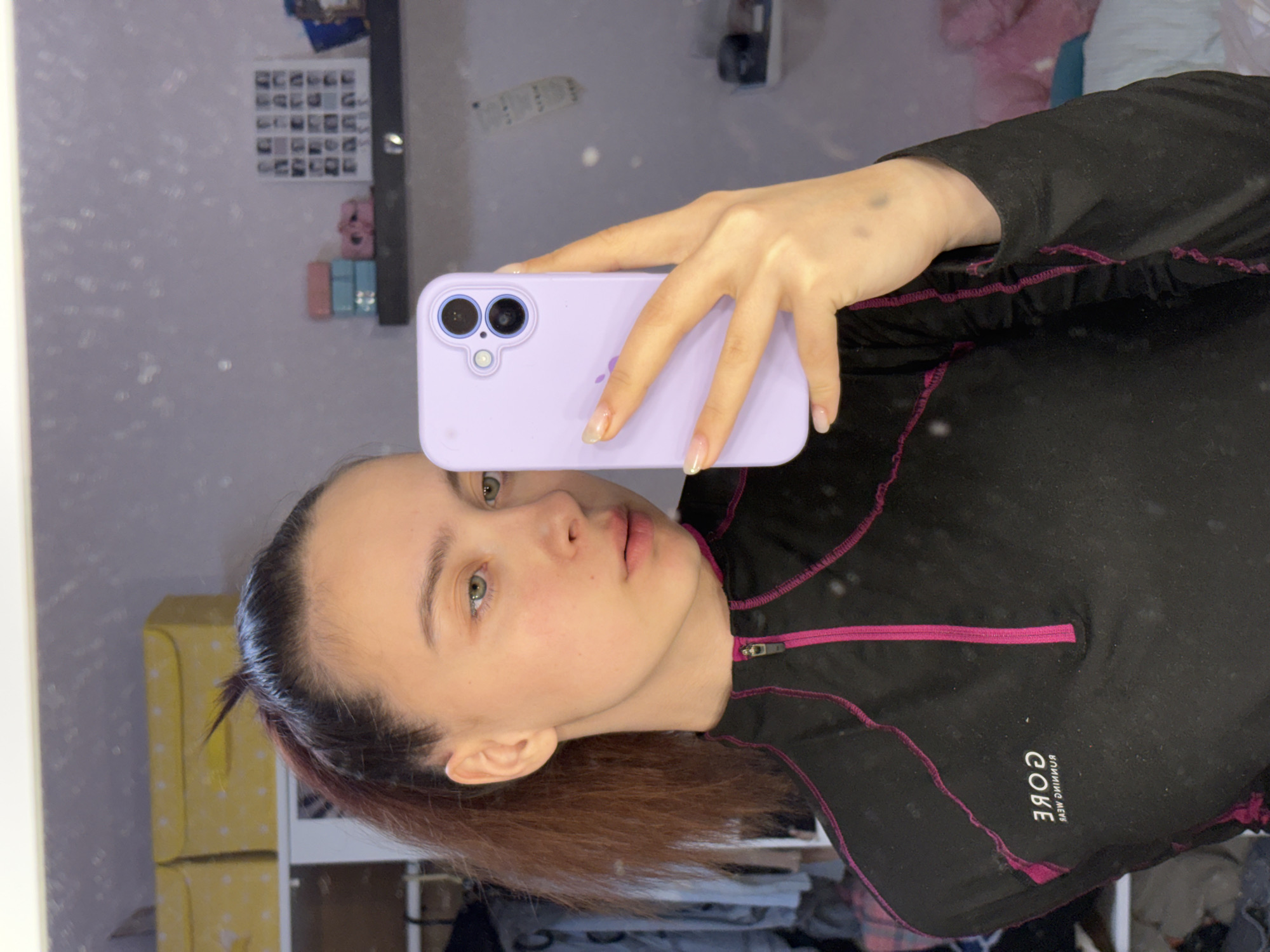 A woman with fair skin and light green eyes is taking a selfie in a mirror. She is holding a light purple iPhone with two visible rear cameras. She is wearing a black athletic jacket with pink trim and the word "CORE" printed on the right side of her chest in a reversed font. The background shows a reflection of a room with items on shelves and posters on the wall. The scene appears to be indoors in Прикамский, Russia.