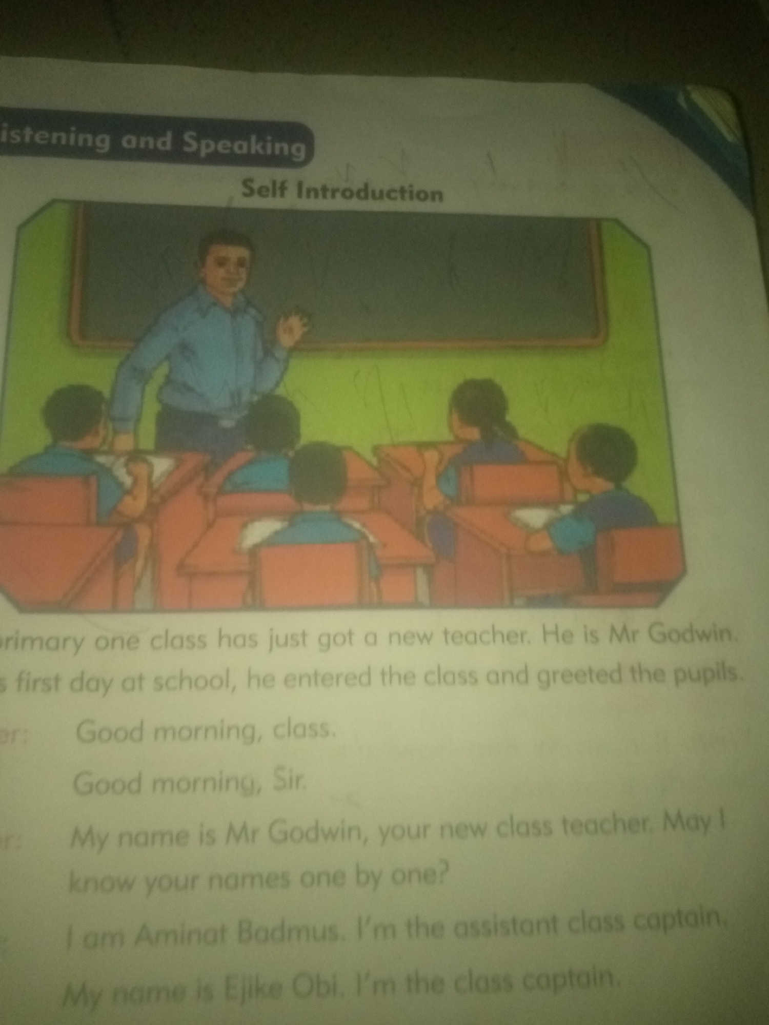 The image depicts an illustration from a children's book, featuring a classroom scene with a teacher and students. A male teacher, dressed in a blue shirt and dark pants, stands at the front of the classroom, facing a group of students seated at desks. The students are also wearing blue shirts. The text below the illustration includes a narrative about a new teacher named Mr. Godwin and some dialogue.
This image is not located in the city of Jauro Yinu, Nigeria. There is no contextual information within the image itself to suggest any specific geographical location, especially not Jauro Yinu. The image is purely illustrative.