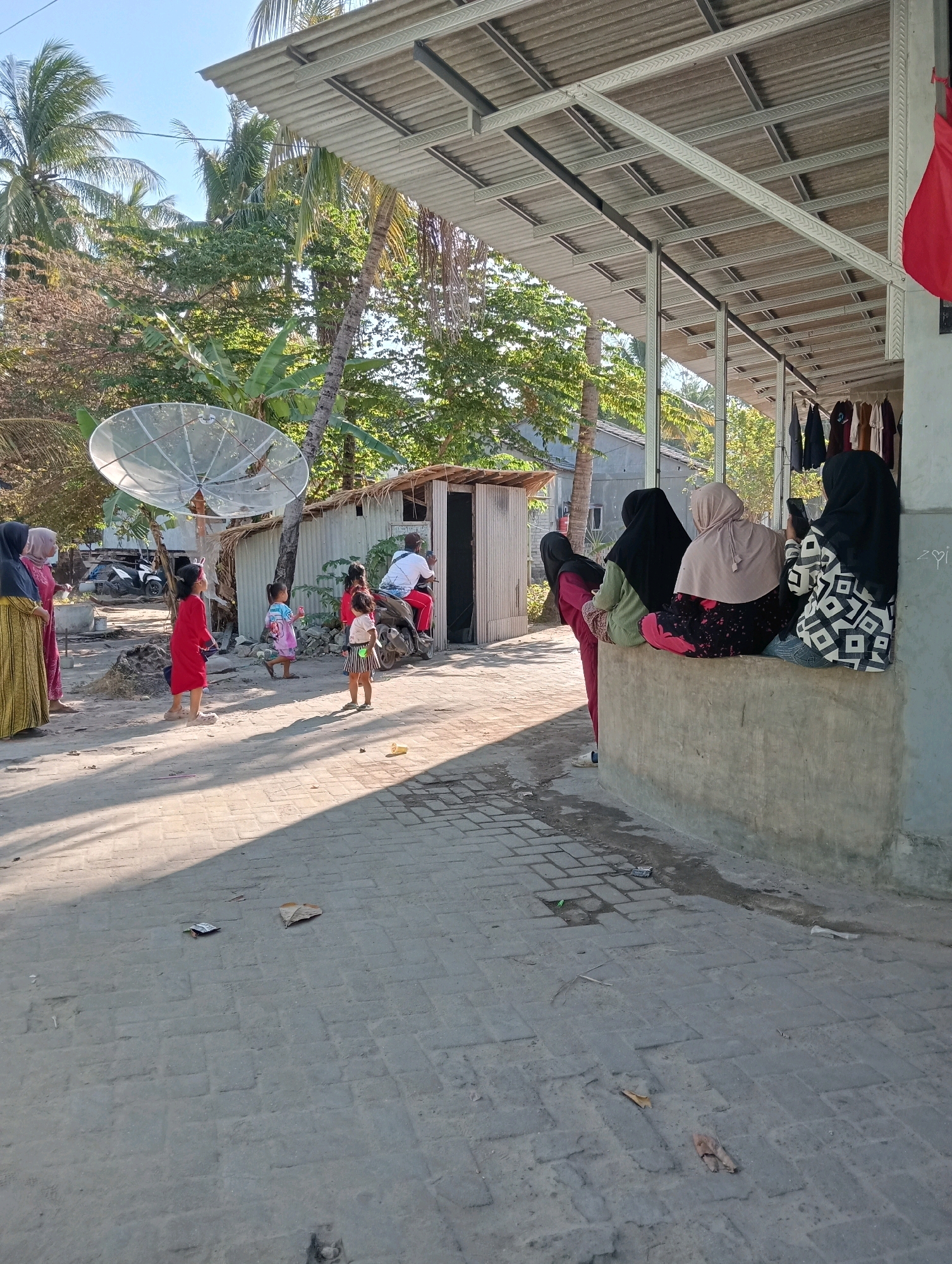 The image depicts a street scene in Pagerungan Kecil, Indonesia, under a clear blue sky. A corrugated metal roof supported by metal beams extends from a building on the right, providing shade over a concrete ledge where five individuals are seated. Three of them are women wearing hijabs, one of whom is looking at her phone. Two children are also present, one girl in a pink dress and another in a striped skirt. On the left, two women in hijabs and traditional dresses stand near a satellite dish. A man on a motorcycle is visible in the background. The ground is paved with interlocking bricks, with some debris and fallen leaves scattered on it. The environment is characterized by tropical vegetation, including palm and banana trees, and a small shed with a corrugated metal exterior.