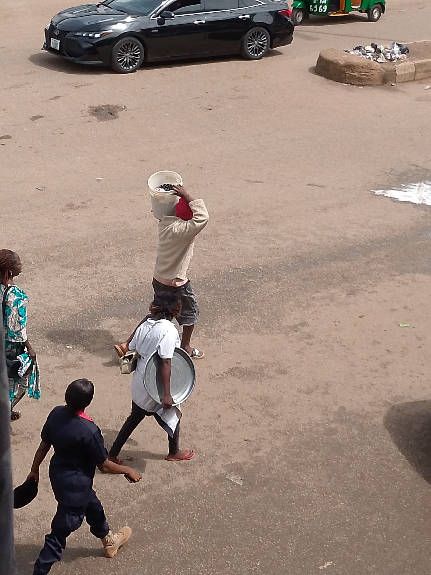 In Bukuru, Nigeria, a black car is parked on a dirt road, facing left. In the foreground, three individuals are walking. A woman in a patterned blue and white dress walks from left to right. Behind her, a person wearing a white shirt and dark shorts carries a bucket of water on their head, with a red object inside. To the right of this person, another individual in a white shirt and dark pants carries a metal tray. Further to the right, a person in a dark blue uniform walks from right to left, carrying a black hat. In the background, a green tricycle with a registration plate reading "PIA 6569" is visible. The scene appears to be an outdoor market or roadside setting.