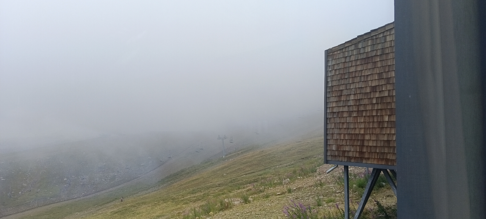 The image shows a foggy mountain landscape. On the right side, there is a modern wooden structure on stilts, possibly a small building or observation deck, with a roof covered in wooden shingles. The structure is partially obscured by a dark grey vertical panel on the far right. The background is filled with dense fog, obscuring most of the landscape. However, faint outlines of a ski lift with multiple chairs can be seen ascending a grassy slope. The ground in the foreground is covered with grass, small rocks, and some purple wildflowers. There are no people clearly visible in the image, though there appear to be two small figures in the distance on the left side of the slope, obscured by the fog. The overall scene suggests a mountainous area, likely with ski resorts, but the weather conditions are poor due to heavy fog. This scene is not in Almaty, Kazakhstan.