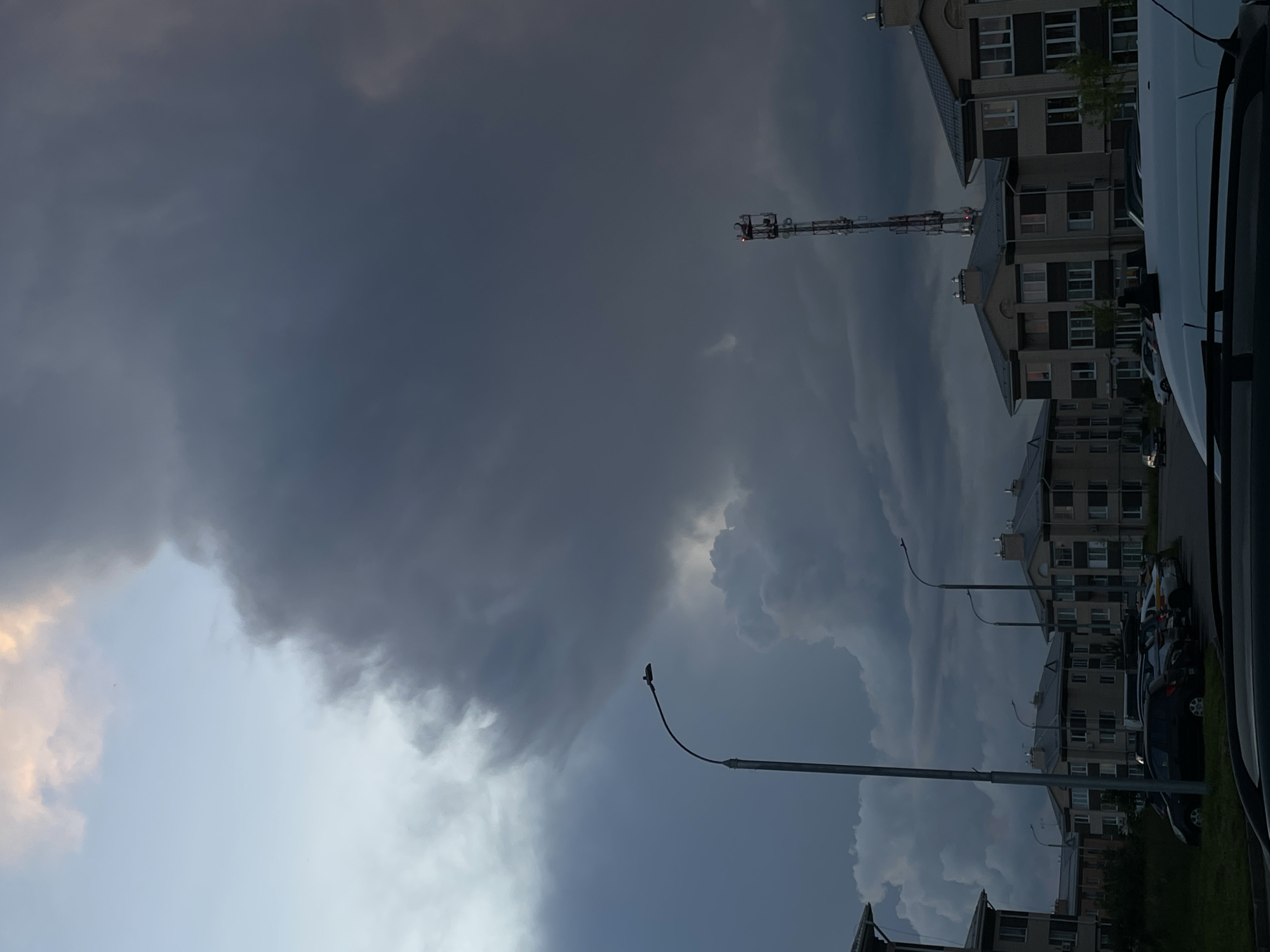 The image depicts a view of a residential area in Zverevo, Russia, under a dramatic, overcast sky. Large, dark storm clouds dominate the upper portion of the frame, contrasting with a sliver of lighter sky with hints of pink and yellow light peeking through on the upper left. In the foreground, several multi-story apartment buildings with light-colored facades and dark roofs are visible. A row of parked cars, including a black sedan and a white van, are situated in front of the buildings. Streetlights with curved poles are positioned along the street. A tall telecommunications tower with antennas stands prominently in the middle ground to the right of the frame. The overall atmosphere is somber, suggesting an impending storm.