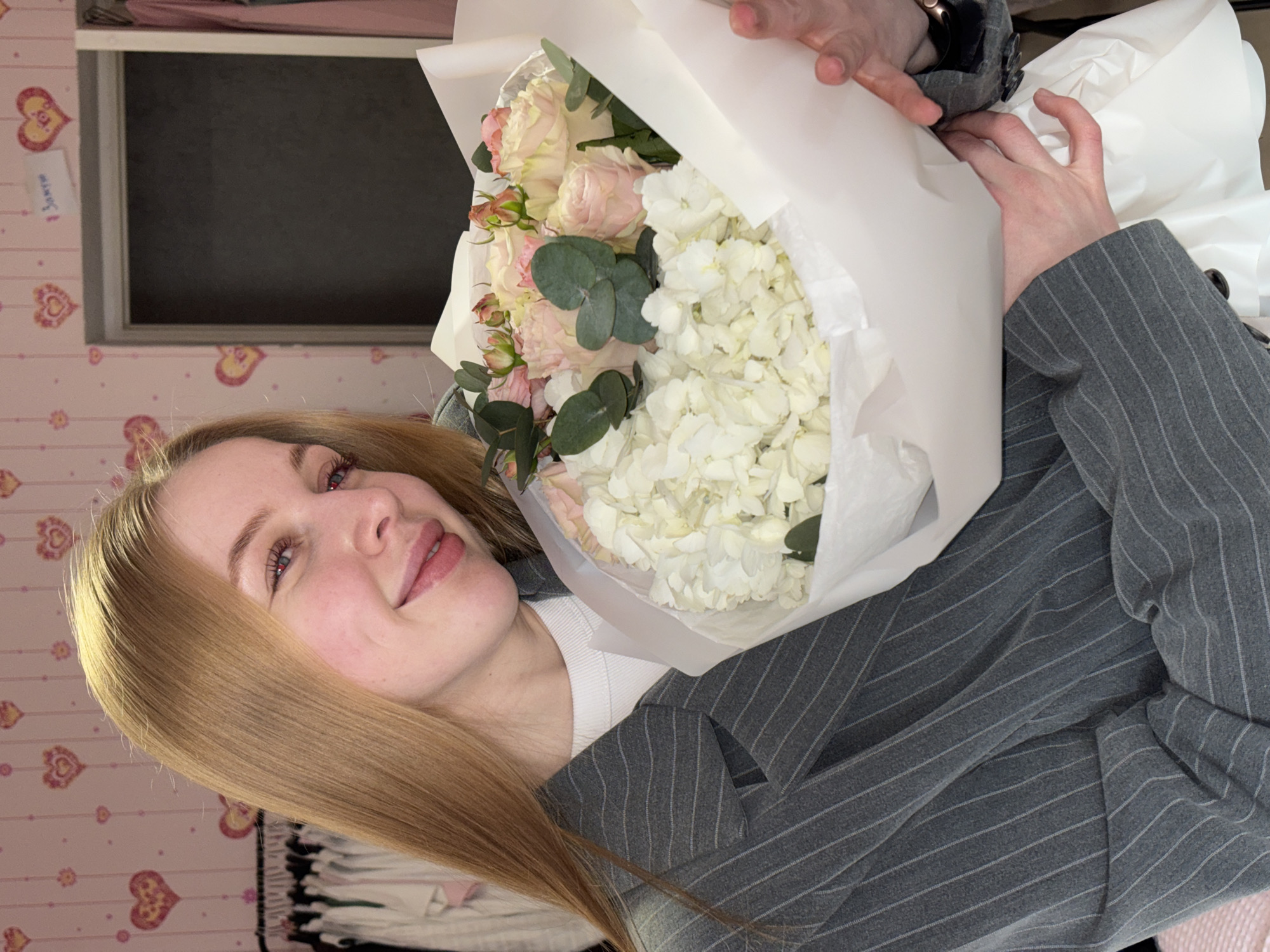 A young woman with long, straight, light brown hair is captured in a close-up, tilted shot, holding a vibrant bouquet of flowers. She is smiling softly, looking towards the camera. Her eyes are light-colored, and she has visible eyelashes. She is wearing a grey pinstripe blazer or jacket over a white top. A metallic smartwatch, possibly an Apple Watch, is visible on her left wrist as her hands delicately adjust the bouquet's wrapping.
The bouquet features a blend of light pink roses (or similar delicate pink flowers), clustered white hydrangeas, and green eucalyptus leaves, all wrapped in pristine white paper.
The background shows a room adorned with pink wallpaper patterned with yellow and orange hearts. In the upper left quadrant of the image, a dark, rectangular window or opening is visible, suggesting an indoor setting. Taped to the heart-patterned wallpaper, next to the window, is a small white note with Russian text that appears to read "3 зачет" (likely meaning "3 credit" or "3 pass," possibly referring to an academic grade or task completion). The overall ambiance is personal and warm, potentially a bedroom or living space in Perm, Russia.