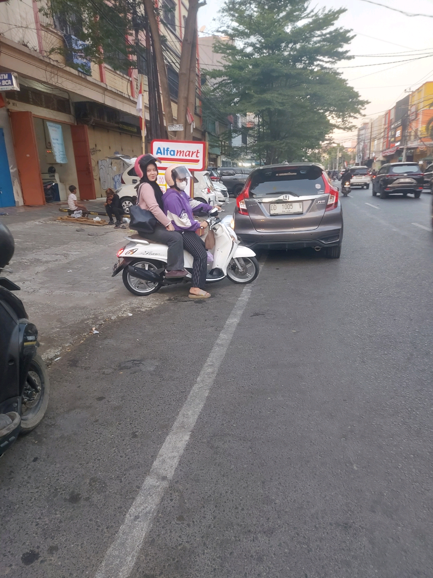 The image shows a street scene in Makassar, Indonesia. Two women are riding a white scooter on the right side of the street. The woman in front is wearing a pink jacket and the woman behind her is wearing a purple jacket and a helmet. They are both looking towards the camera. In the background, there are buildings, trees, and other vehicles, including cars and motorcycles. A sign for "Alfamart" is visible on the left side of the street. The road is asphalt and has a white line marking the lane. The overall scene depicts a typical urban environment with moderate traffic.