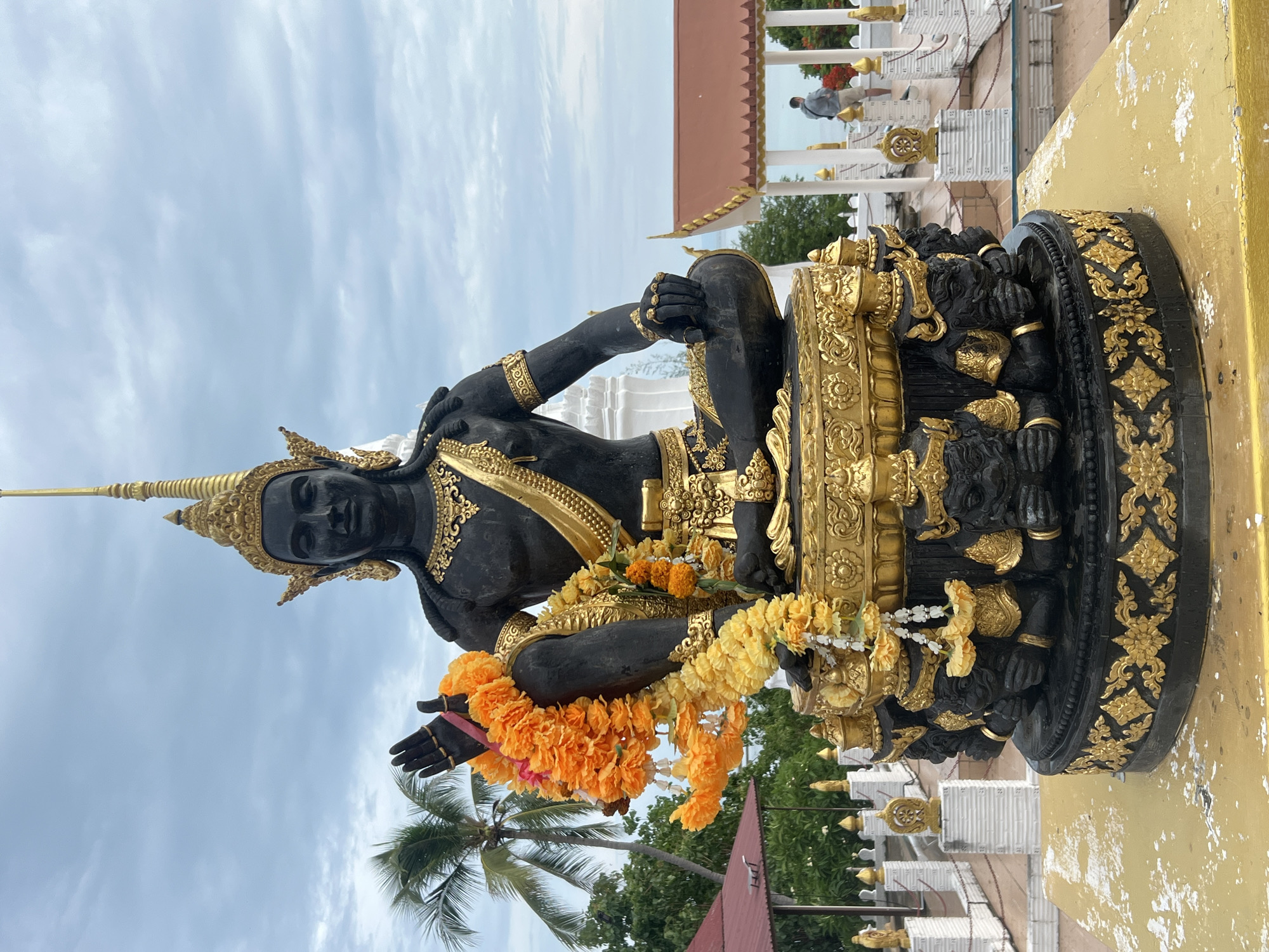 The image is a full shot of a black statue adorned with gold jewelry and garlands of orange and yellow flowers. The statue sits cross-legged on a gold-embellished pedestal. In the background, there's a white structure, possibly part of a temple or religious site, and a glimpse of a man standing on a raised platform. Palm trees and other greenery are visible, suggesting an outdoor location. The sky is overcast with patches of blue. Based on the image title, the location is Baan Hua Thanon, Thailand.