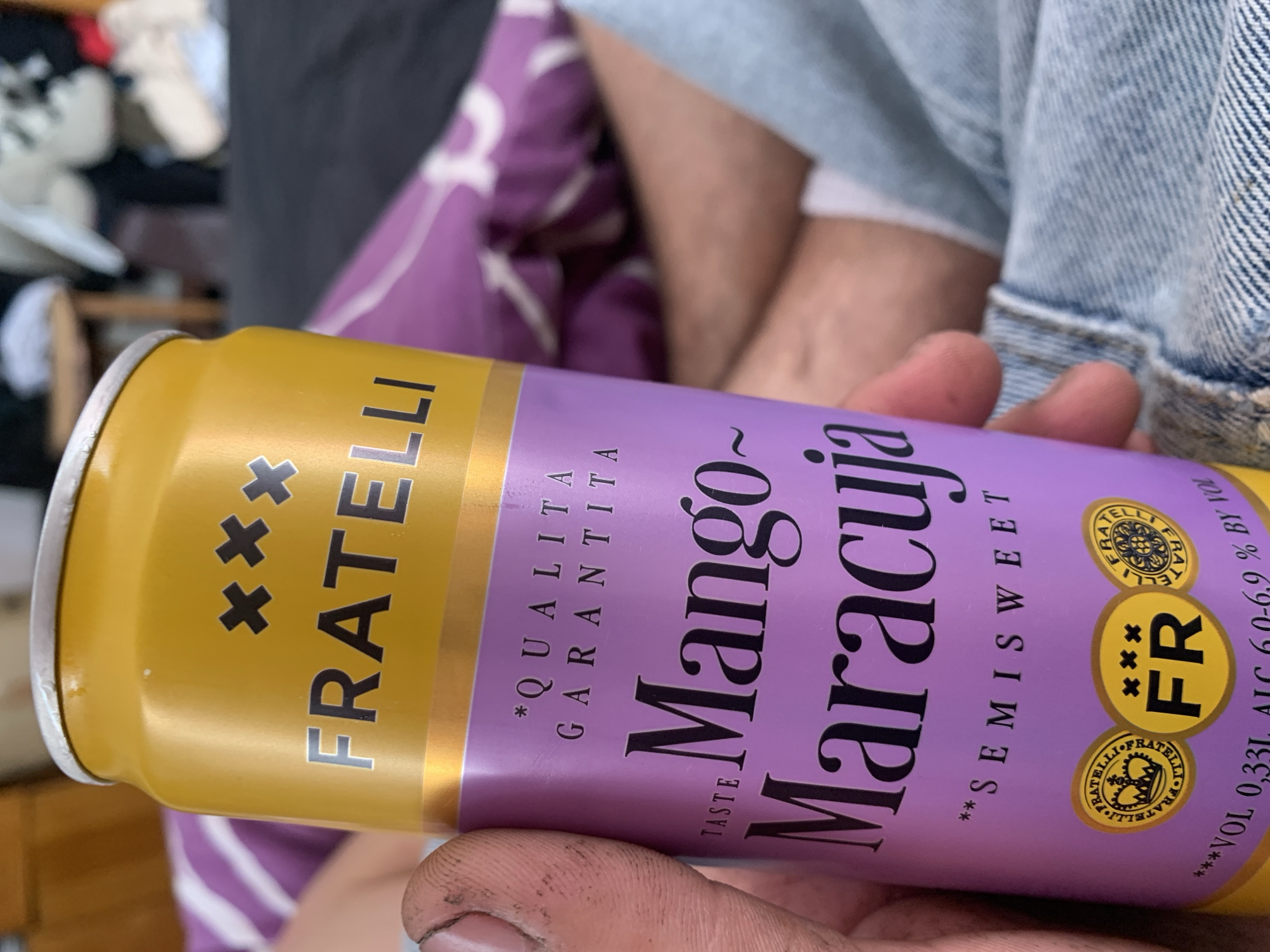 The image shows a close-up of a person's hand holding a can of "FRATELLI Mango Maracuja Semisweet" beverage. The can is predominantly yellow and purple, with the brand name "FRATELLI" in bold black lettering on the yellow upper portion. The purple section displays text such as "*QUALITA GARANTITA," "TASTE Mango~Maracuja," and "SEMISWEET." At the bottom of the can, there are round emblems and text indicating "VOL 0,33L ALC 6,0-6,9 % BY VOL". The background is blurred, but appears to be an indoor setting with a purple and white striped surface, possibly bedding, and some clothing in the distance. The image does not contain any elements that would allow for the determination of its location within the city of Zhmerynka, Ukraine, or any other specific geographical location.