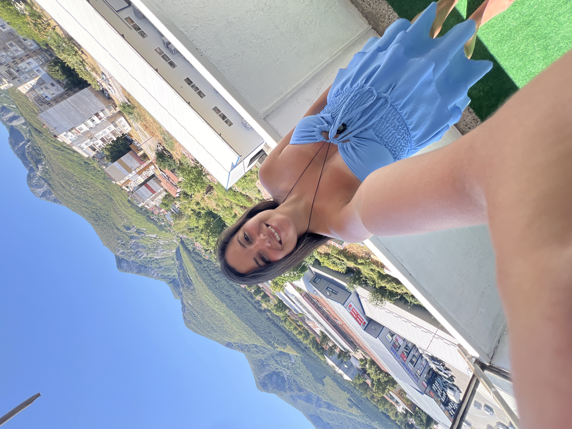 A woman is taking a selfie from a balcony in Vratsa, Bulgaria. She is wearing a blue, strapless dress with ruffles and a necklace. Behind her, the city of Vratsa is visible, with buildings nestled against a backdrop of lush, green mountains under a clear blue sky. The balcony has a railing and a patch of artificial turf.