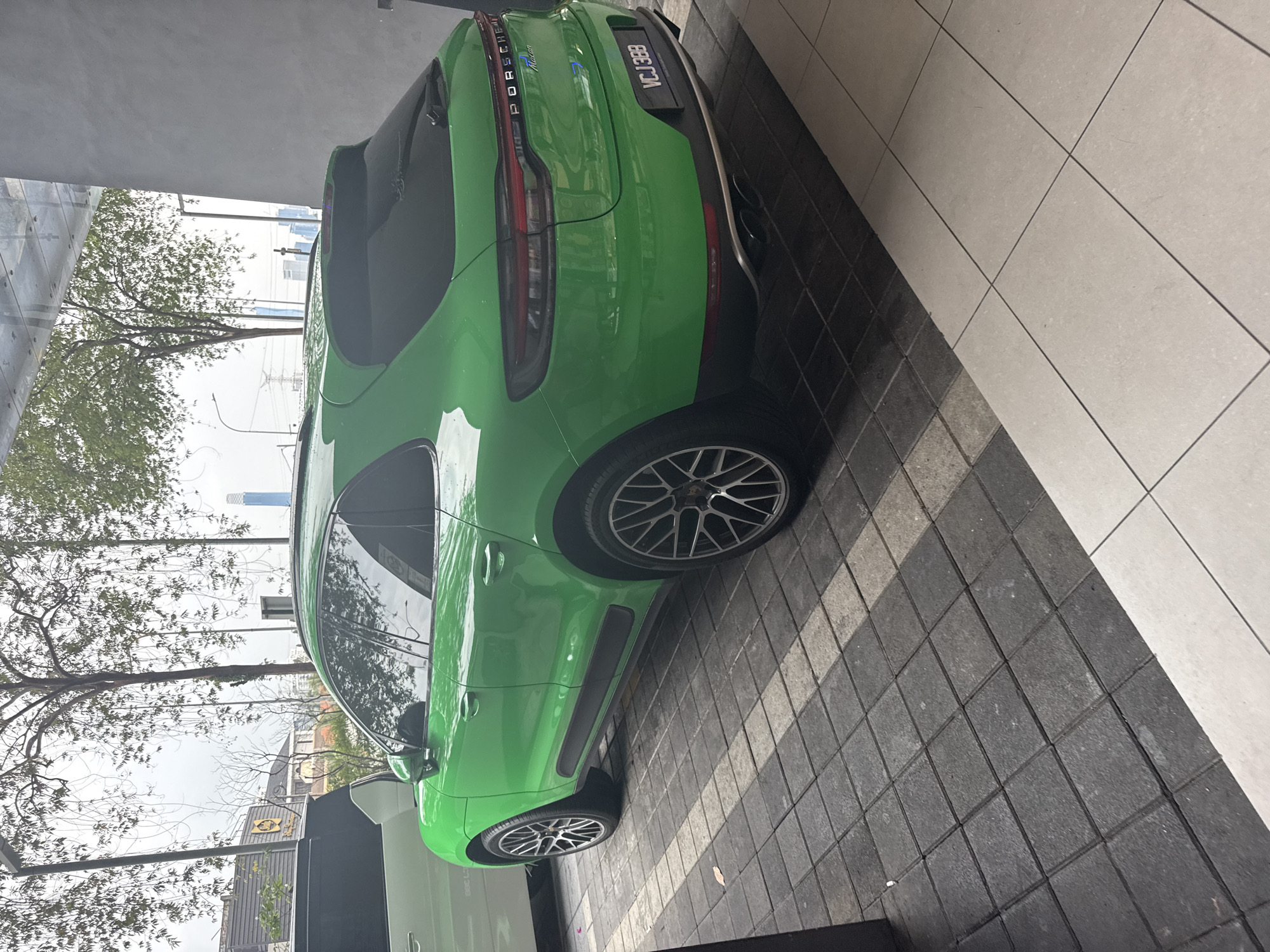 The image shows a green Porsche Macan SUV parked on a grey tiled surface. The vehicle is parked next to a grey wall with a tree visible in the background, along with a partial view of buildings. The license plate, visible on the car's rear, reads "VCJ 3888". Based on the presence of Malaysian license plate and buildings matching similar structures in Kuala Lumpur, it's probably located in Kuala Lumpur, Malaysia. The surroundings suggest an urban setting.