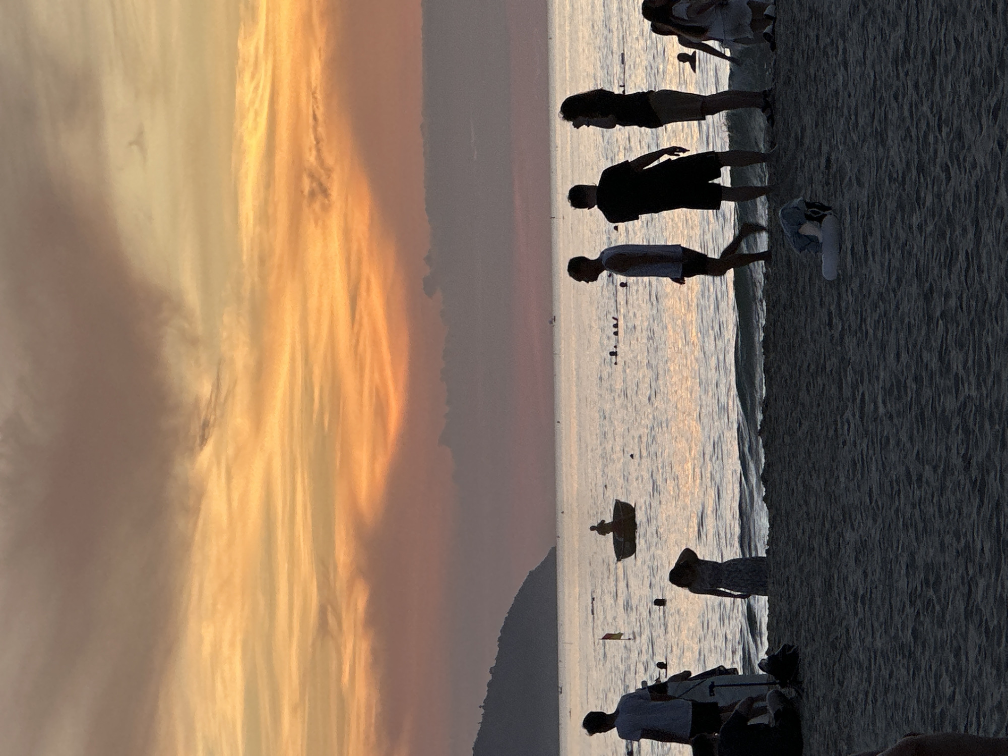 The image depicts a sunset scene at My Khe Beach in Da Nang, Vietnam. Silhouetted figures of people are visible on the sandy beach and in the shallow water. In the foreground, several individuals are standing and walking, with one person kneeling on the sand. Further out in the water, more people are seen swimming or wading. A small boat with a person inside is positioned in the middle ground, near a red and yellow flag. The ocean stretches to the horizon, where it meets a hazy sky filled with vibrant orange, yellow, and pink clouds, characteristic of a sunset. A dark, mountainous landmass is visible on the left side of the horizon. The overall atmosphere is peaceful and serene, with the warm hues of the sunset casting a glow over the scene.