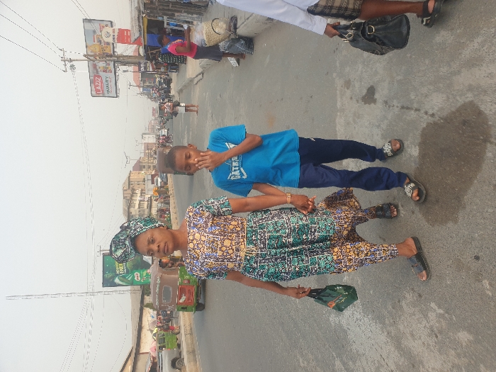 On a busy street, a woman in a patterned dress and headwrap holds hands with a young boy in a blue shirt and navy pants. Behind them are vehicles and buildings, including a billboard advertising Laziz. The woman carries a small bag, and the boy has his hand to his face. A few other people can be seen in the background, including one carrying a purse.