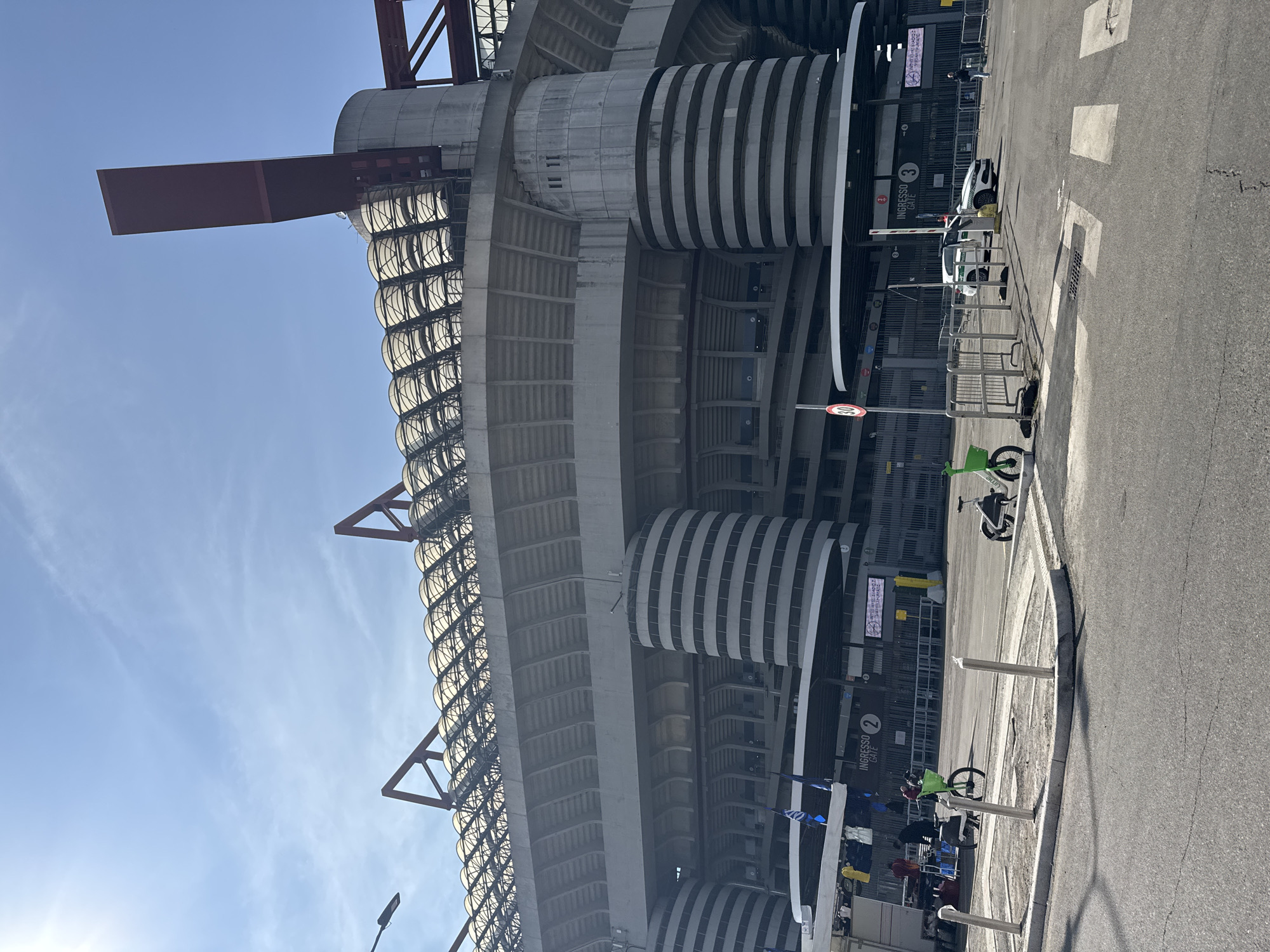 The image shows the San Siro Stadium in Milan, Italy, from an exterior ground-level perspective. The stadium's characteristic concrete structure and spiraling striped towers are prominent. A clear, blue sky is visible above. The foreground includes a street, sidewalk, parked scooters, and crowd control barriers. The light suggests it's daytime with possible direct sunlight. There appear to be a few people near the entrance gates.