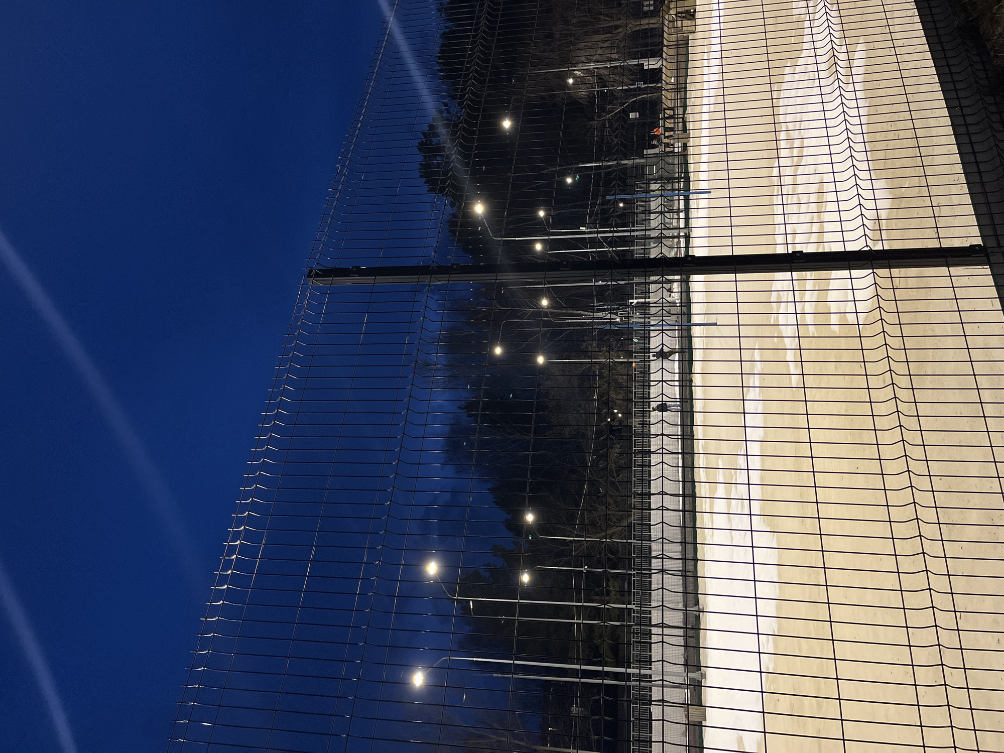 The image shows a view through a chain-link fence at night, likely in Chaikovsky, Russia. On one side of the fence is a dark blue sky. On the other side, behind the fence, are trees, streetlights, and what appears to be a building or structure. The streetlights cast a warm glow, while the building has a white facade.