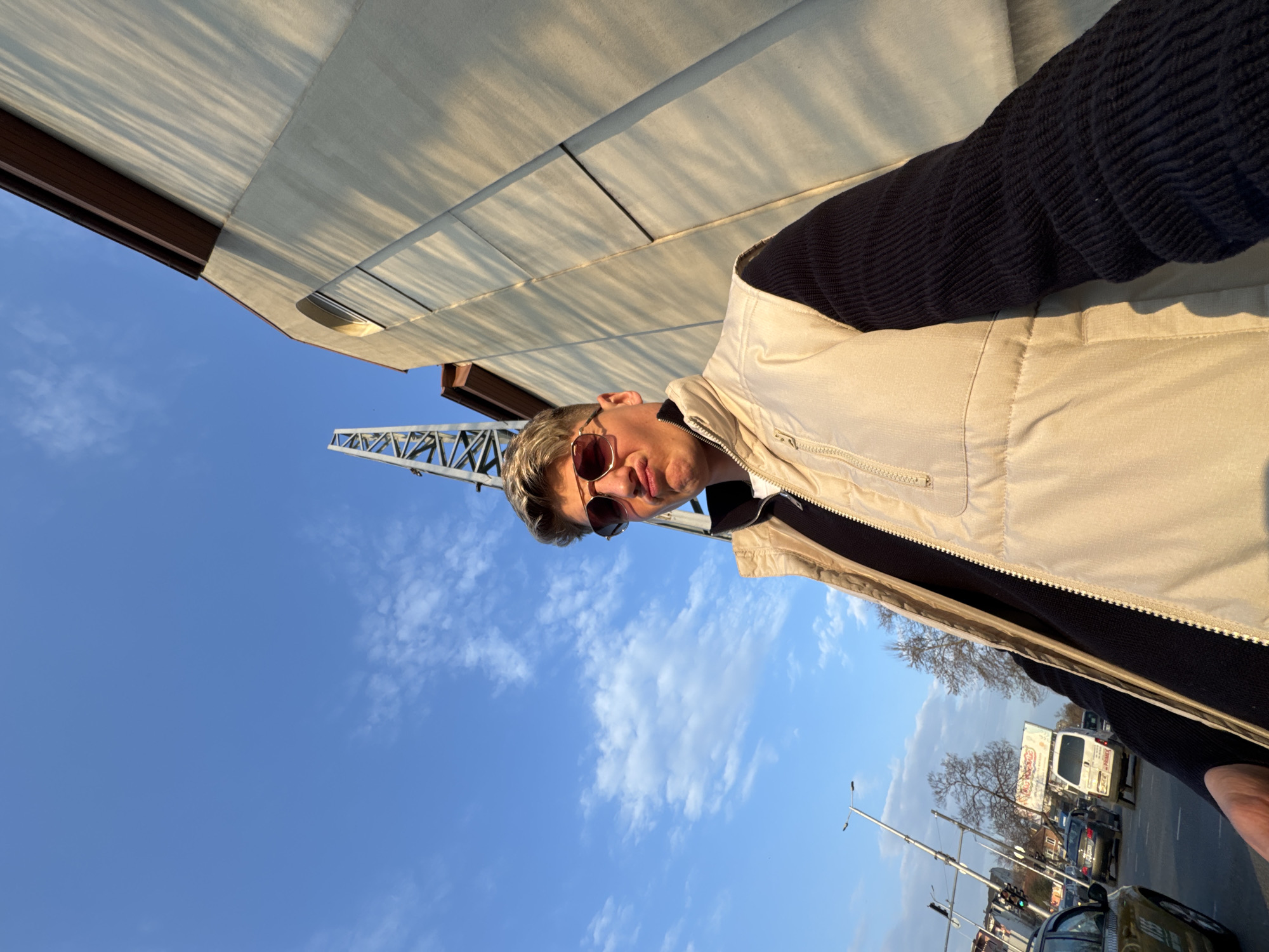 A young male, possibly in his late teens or early twenties, is depicted in a low-angle selfie, prominently filling the left and center foreground. He faces the camera, wearing aviator-style sunglasses with reddish-brown lenses and exhibiting a playful pouting expression. His hair is light-colored and styled upwards. He is dressed in a light beige quilted vest over a dark, long-sleeved ribbed or corduroy shirt. His left hand is partially visible in the lower right, holding the camera.
The scene is an urban outdoor setting under a clear blue sky with scattered white clouds. Strong direct sunlight from the right casts defined shadows. To the immediate right and extending upwards, a large, light-colored (beige/off-white) building with a brown roofline is visible, casting long, diagonal shadows across its facade, indicating a low sun angle. A semi-circular arched window or architectural feature is visible on the upper part of the building.
To the left and in the background, a busy street is active with several vehicles, including a light-colored van and a yellow taxi. Traffic lights with an illuminated green signal are visible on a pole. Further down the street, a large billboard with Cyrillic script and leafless trees are present. A tall, triangular metal lattice structure, possibly a crane or a supporting mast, rises vertically behind the subject and the streetscape, to the left of the building.
The location is Plovdiv, Bulgaria, specifically in the vicinity of the International Fair Plovdiv complex, identifiable by the distinctive metal lattice structure and the architecture of the adjacent building.