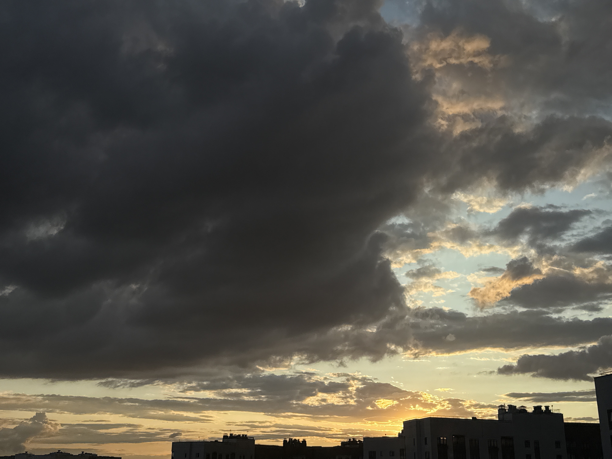 The image depicts a dramatic sky filled with dark, heavy clouds overhead, contrasting with patches of lighter clouds illuminated by a setting or rising sun towards the horizon. The lower portion of the image shows the silhouetted rooftops of buildings in the city of Tomilino, Russia. The architecture appears modern, with flat roofs and visible ventilation structures. The lighting suggests either dawn or dusk, casting a warm, golden glow on the lower cloud layers and creating shadows on the buildings. There are no people or visible actions in the scene, only the natural elements of the sky and the urban landscape.