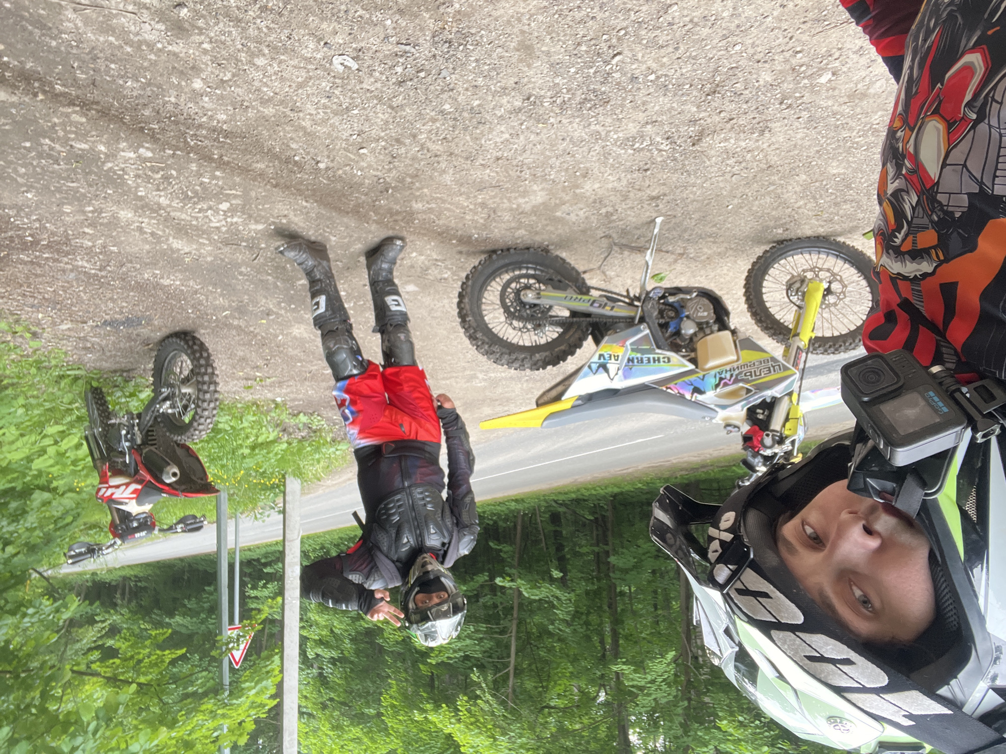 The image captures two individuals in motocross attire on a dirt path alongside an asphalt road in the city of Ореховка, Russia. The scene is bordered by dense green trees and undergrowth, suggesting a wooded or forest environment.
In the immediate foreground on the left, one individual is partially visible, appearing to be taking a selfie. This person wears a white, black, and lime green motocross helmet adorned with "100%" branding on the strap and visor, and a GoPro camera is mounted on the front of the helmet. Their eyes are directed towards the camera. The visible part of their clothing includes a red and black long-sleeved motocross jersey with intricate graphic designs.
To the right and slightly further back, a second individual stands on the dirt path, facing the camera and making a peace sign with their left hand. This person is equipped with a black full-face helmet with goggles, a black protective chest and arm armor worn over a dark garment, red motocross pants featuring white and blue accents, black knee pads, and black motocross boots marked with a white "E" symbol.
Two dirt bikes are present. One, positioned in the midground to the left of the second person, is primarily white, yellow, and black, displaying text such as "ЦЕЛЬ ВЕРШИНА" and "CHERN AEV" on its side panel. It is parked on a kickstand on the dirt. The second dirt bike, red and white with "JMC" branding visible on its handlebars, is situated on the far right, partially obscured by foliage, also parked on the dirt path.
In the background, a narrow asphalt road runs parallel to the dirt path. A light-colored utility pole and a red-bordered white triangular warning sign stand beside the road. The ground consists of varied dirt and gravel. The overall lighting suggests an overcast sky.