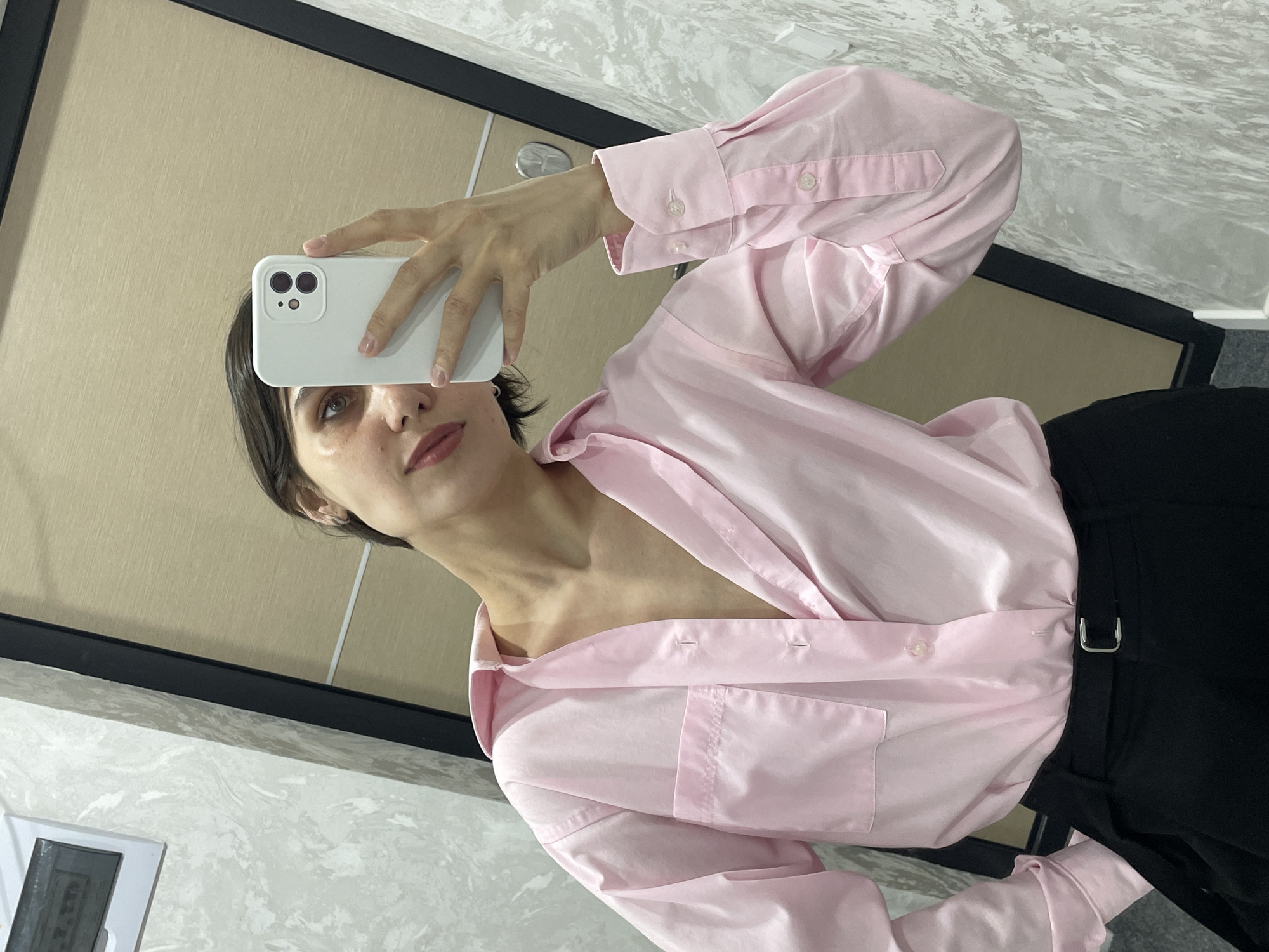 A woman is taking a selfie in what appears to be a room with a mirror. She is holding a white smartphone in her right hand, obscuring part of her face. She has dark hair, fair skin, and is wearing a pink button-down shirt and black pants with a belt. The background consists of a mirror framed in black with a beige wall section visible, and a white wall with a marble-like pattern. A portion of a frame is visible in the lower left corner. It is not possible to tell the location.