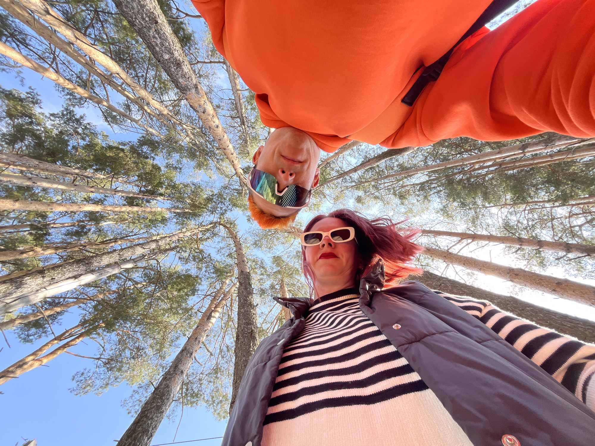 This media file captures two individuals in a low-angle, wide-lens shot looking up into a forest canopy under a bright blue sky. The setting appears to be a coniferous forest, consistent with the provided location context of Chaikovsky, Russia.
One person is prominently featured at the top of the frame, appearing upside down. They have bright orange hair and are wearing a vibrant orange hooded sweatshirt. Their face is adorned with multi-colored, highly reflective, futuristic-style sunglasses, and they display a subtle smile. Below and to the right, the second individual is seen from a more conventional upright perspective. This person has dark reddish-brown hair, is wearing white rectangular sunglasses, and has dark red lipstick. Their attire consists of a black and white striped long-sleeved shirt paired with a dark-colored puffer vest.
The background is filled with numerous tall trees, likely pines or firs, whose straight, light-colored trunks extend vertically into the frame. Patches of green foliage are visible against the clear blue sky, suggesting a bright and sunny day. The overall impression is that of a playful outdoor portrait, likely a selfie taken from a low vantage point, emphasizing the height of the trees and the vastness of the sky. No visible text is present in the image.