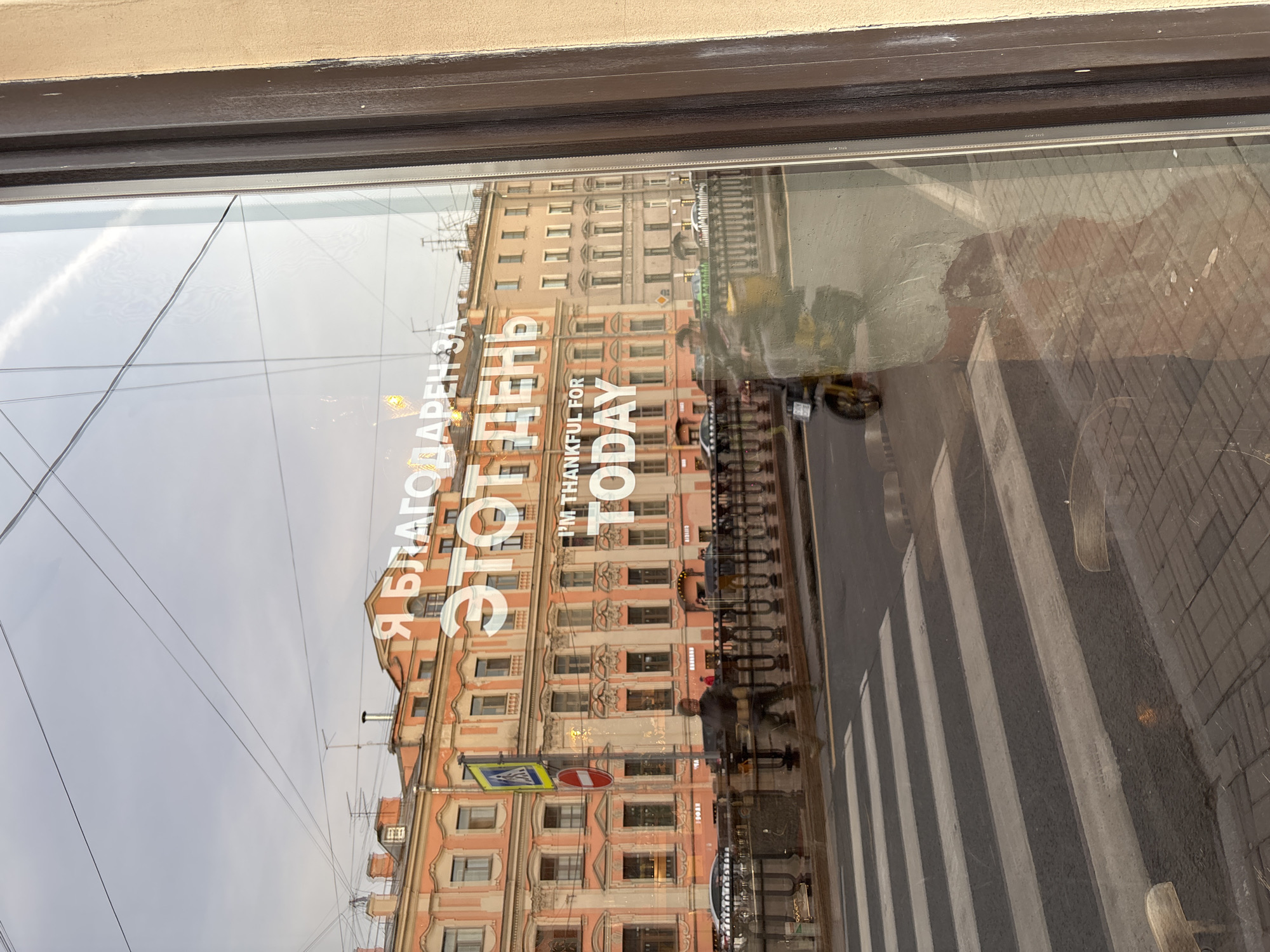 The image shows a window reflection of a building in Saint Petersburg, Russia. The building is multi-storied, with a peach-colored facade. Reflected in the window is a zebra crossing on a street, with people crossing. The text "Я благодарен за этот день / I'M THANKFUL FOR TODAY" is also visible on the window.